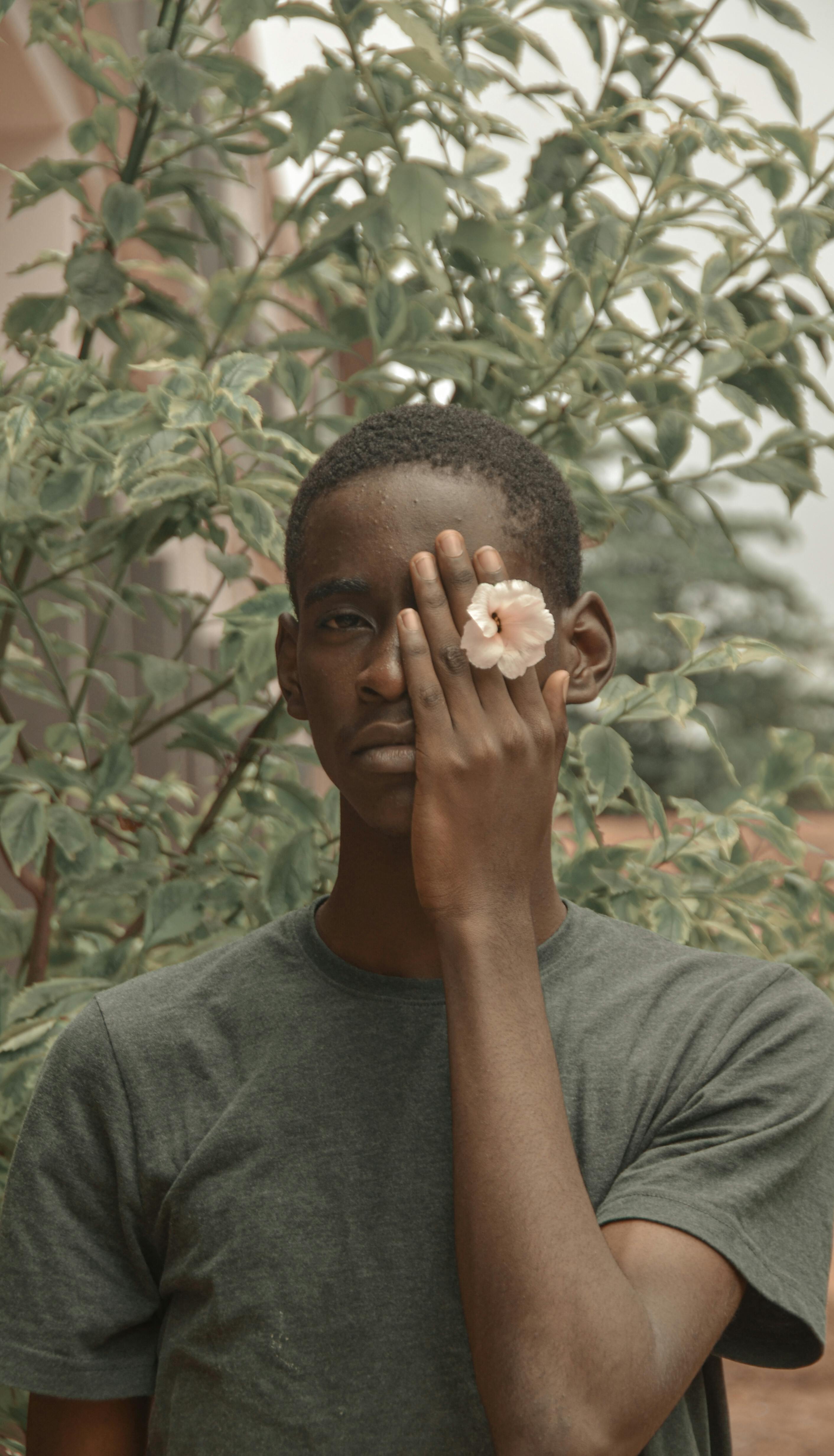 A young man in a gray shirt holds a flower to his face, standing against a leafy backdrop.