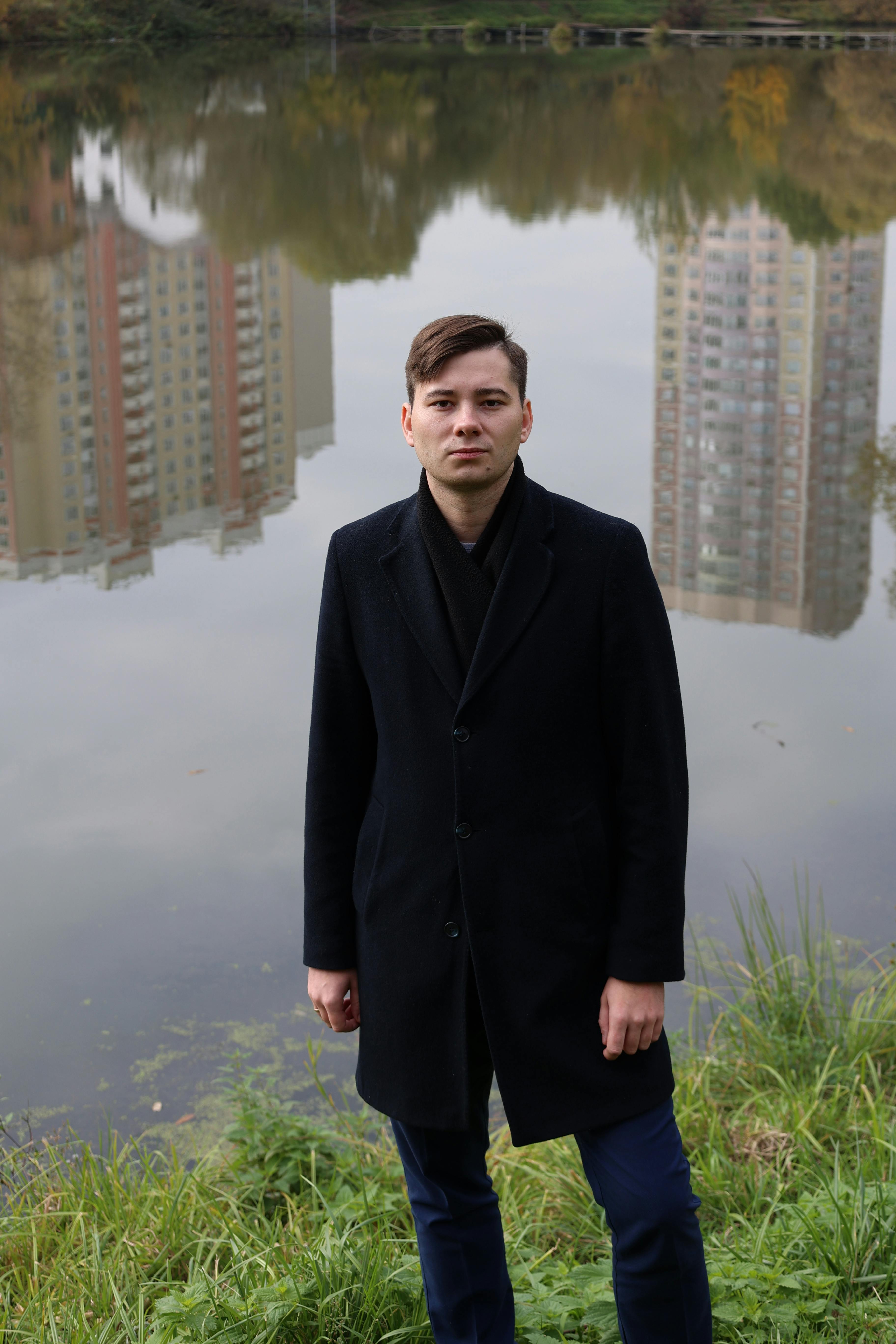 A young man in a black coat stands beside a pond with city reflections.