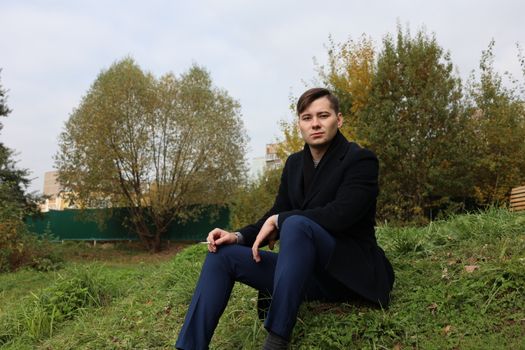 A stylish young man sits outdoors in a park, surrounded by autumn foliage.