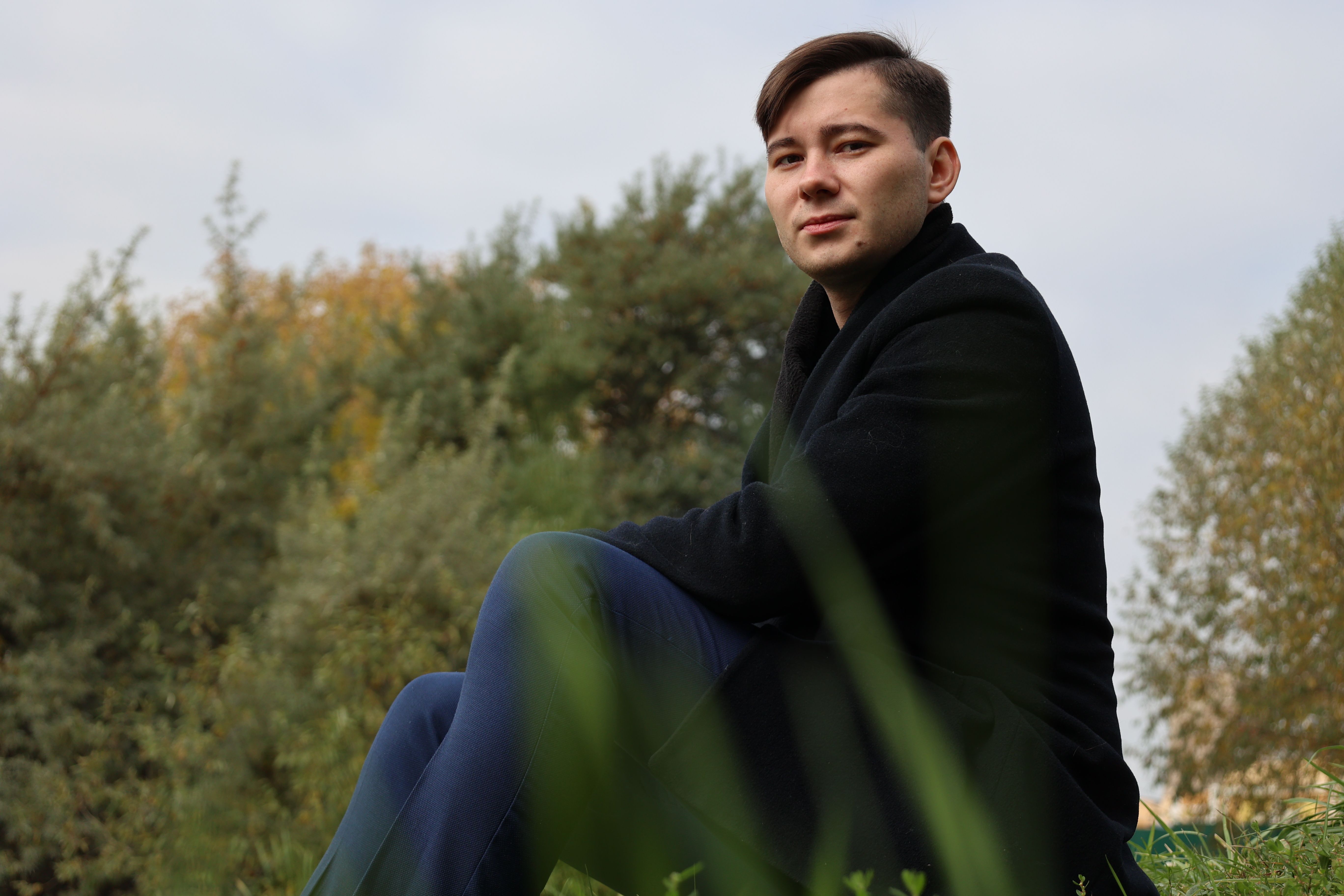 A man sitting in a black coat on grass with trees in the background, embracing nature.