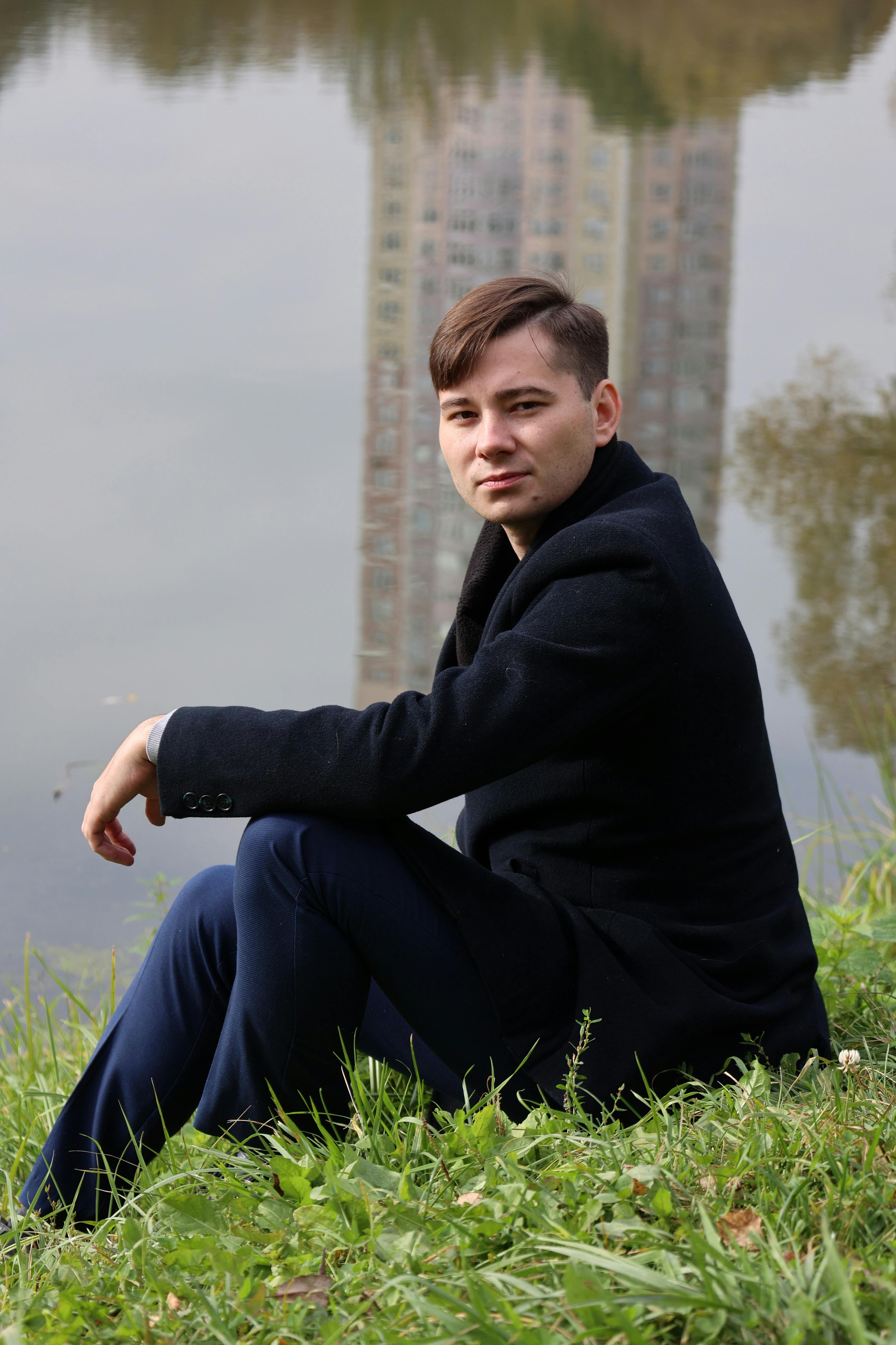 Stylish young man sitting on grass near water reflecting a tall building.