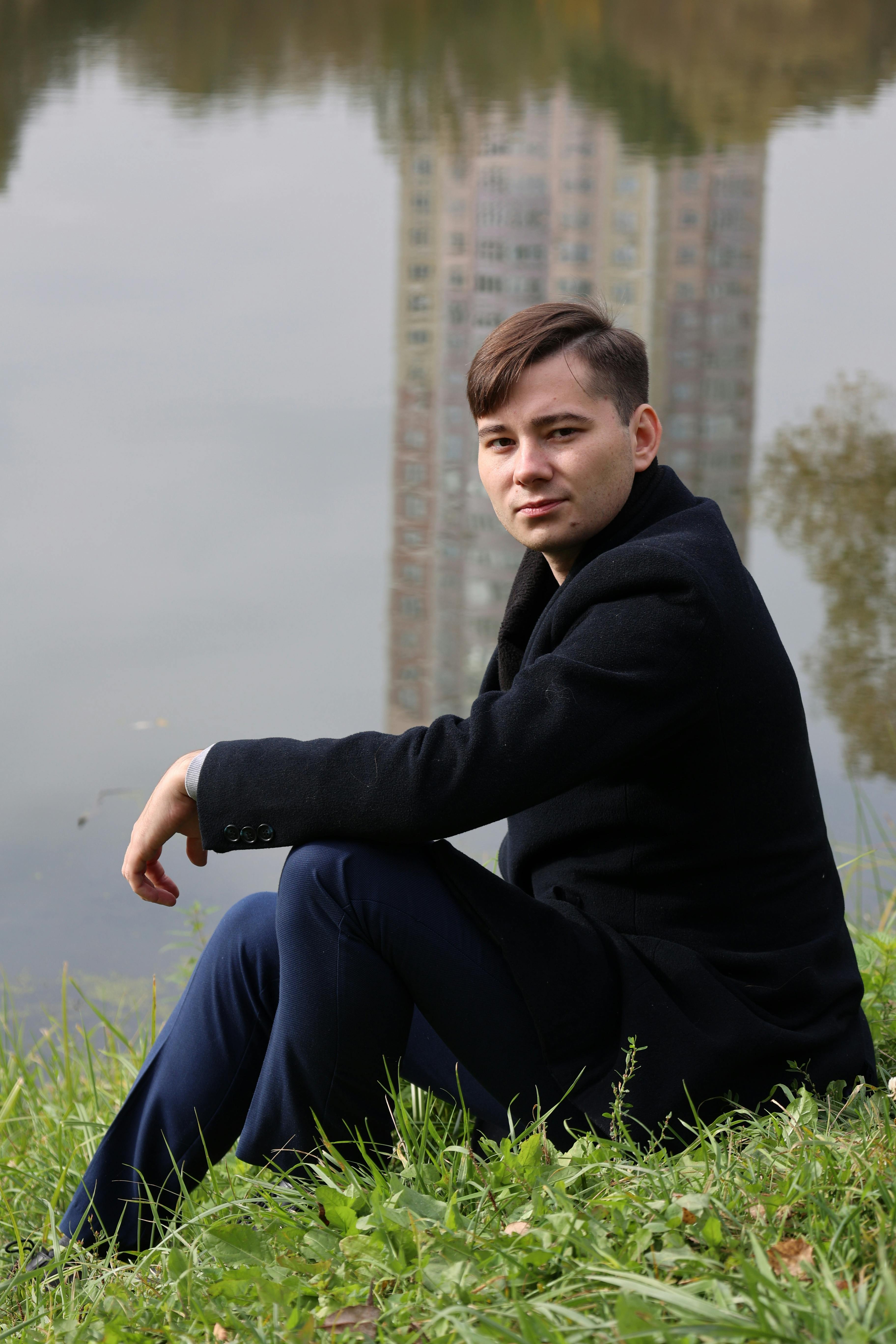 Young man in black jacket sitting by a reflective water scene.