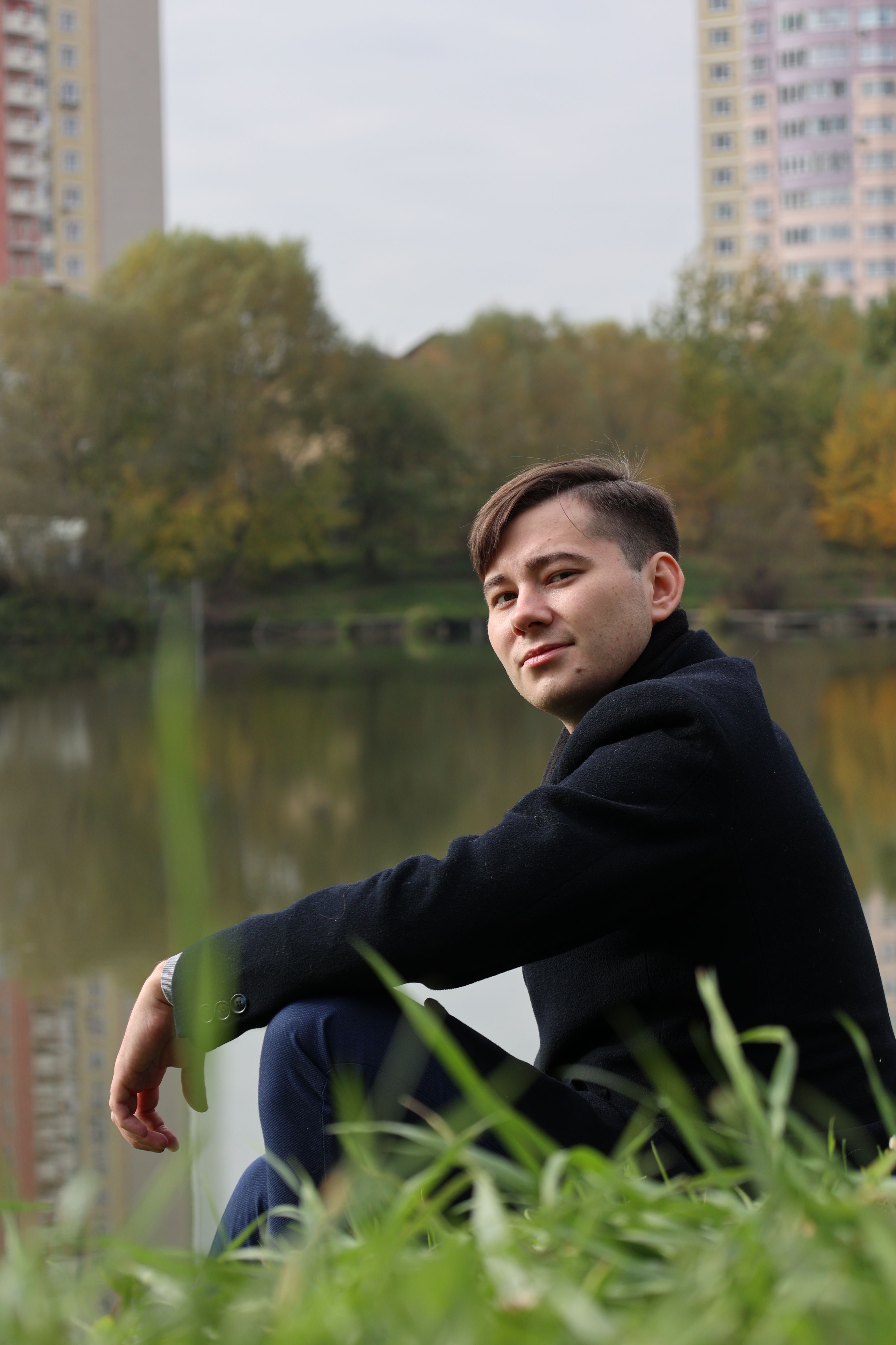 A young man in a black coat sits by a serene urban lake with autumn foliage and city buildings.