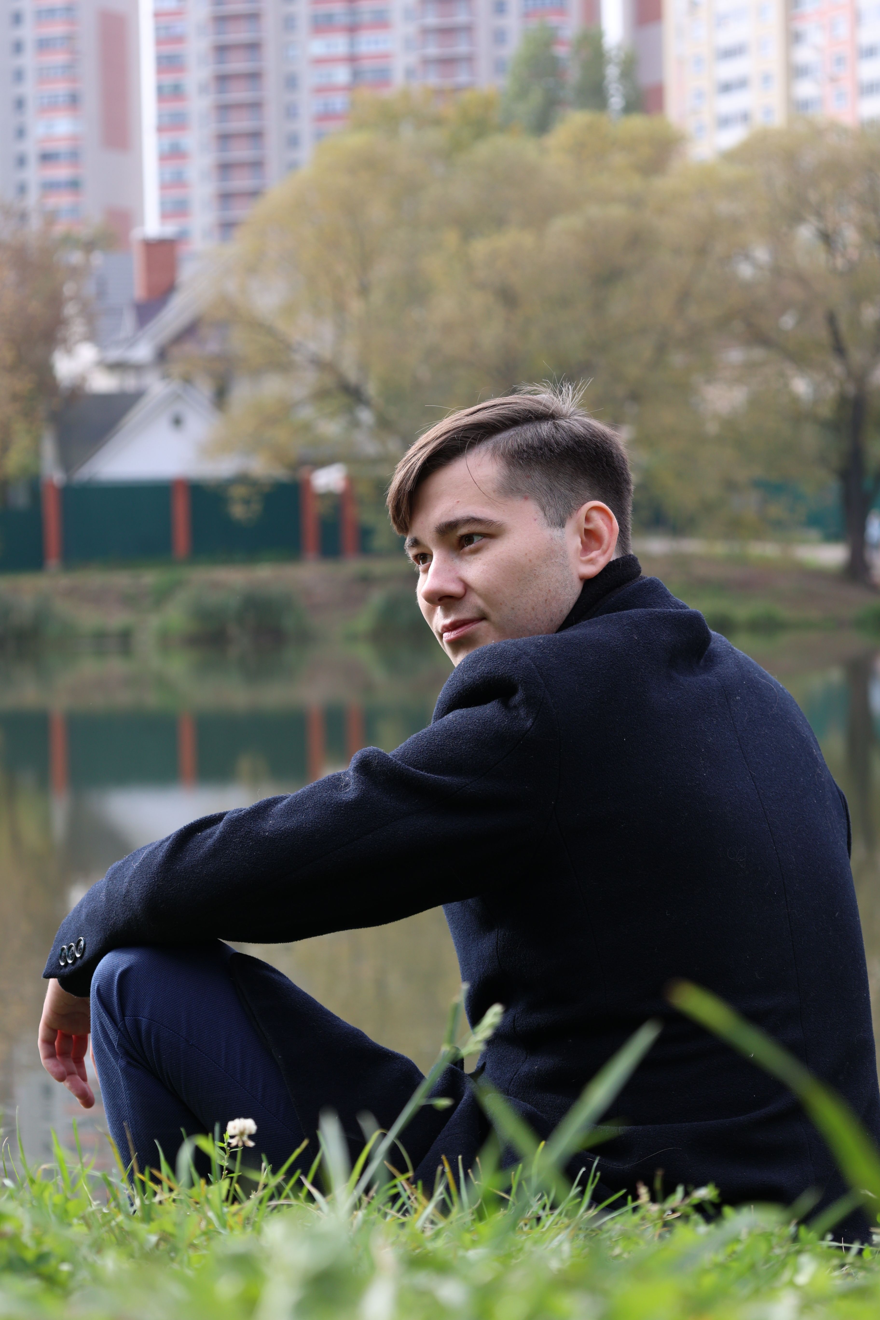 A man in a suit jacket sits by a lake with city buildings in the background.
