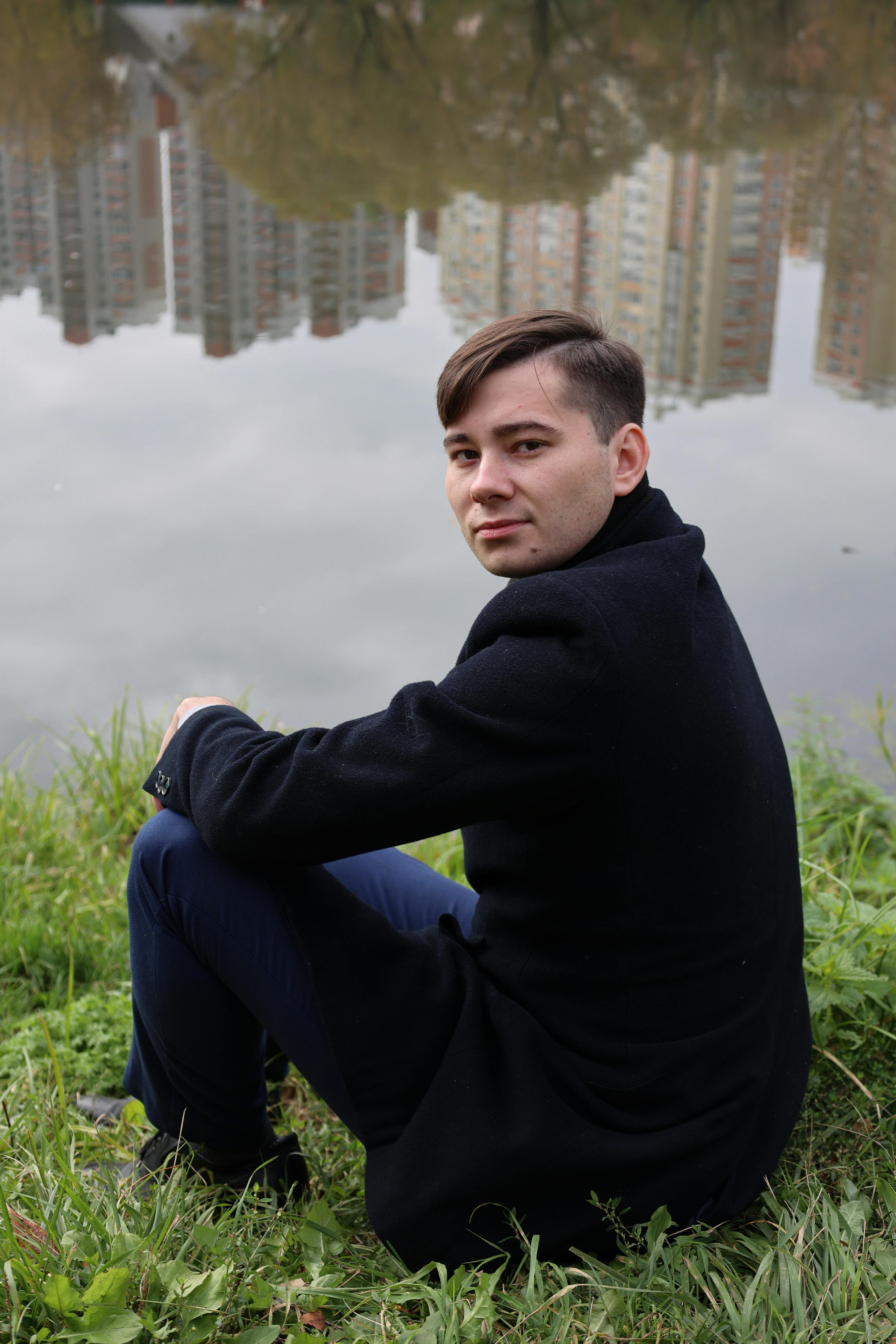 Man in dark coat sits by a river with tall buildings in background, contemplative mood.