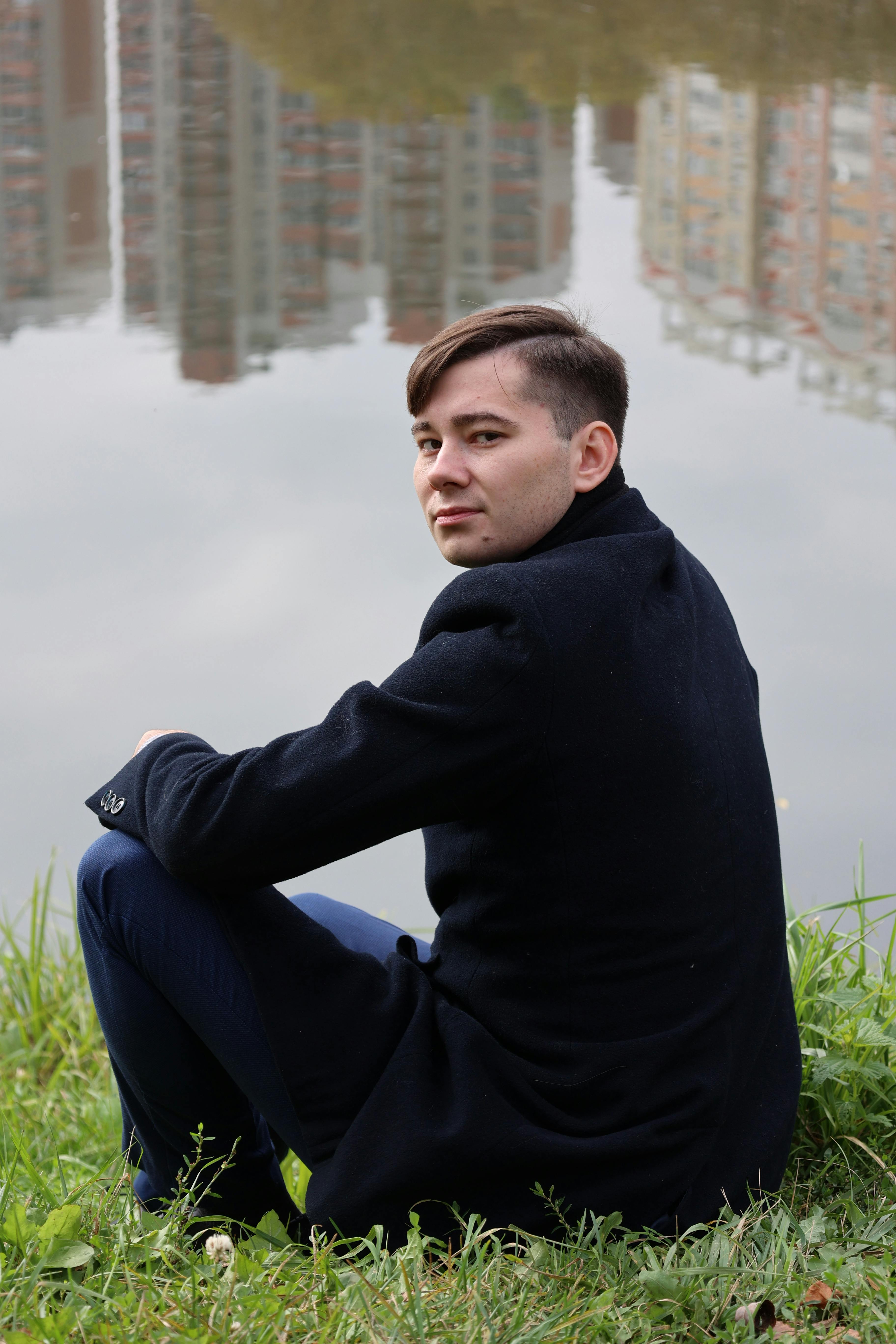 A man in a black coat sits by a lake with city buildings reflecting in the water.