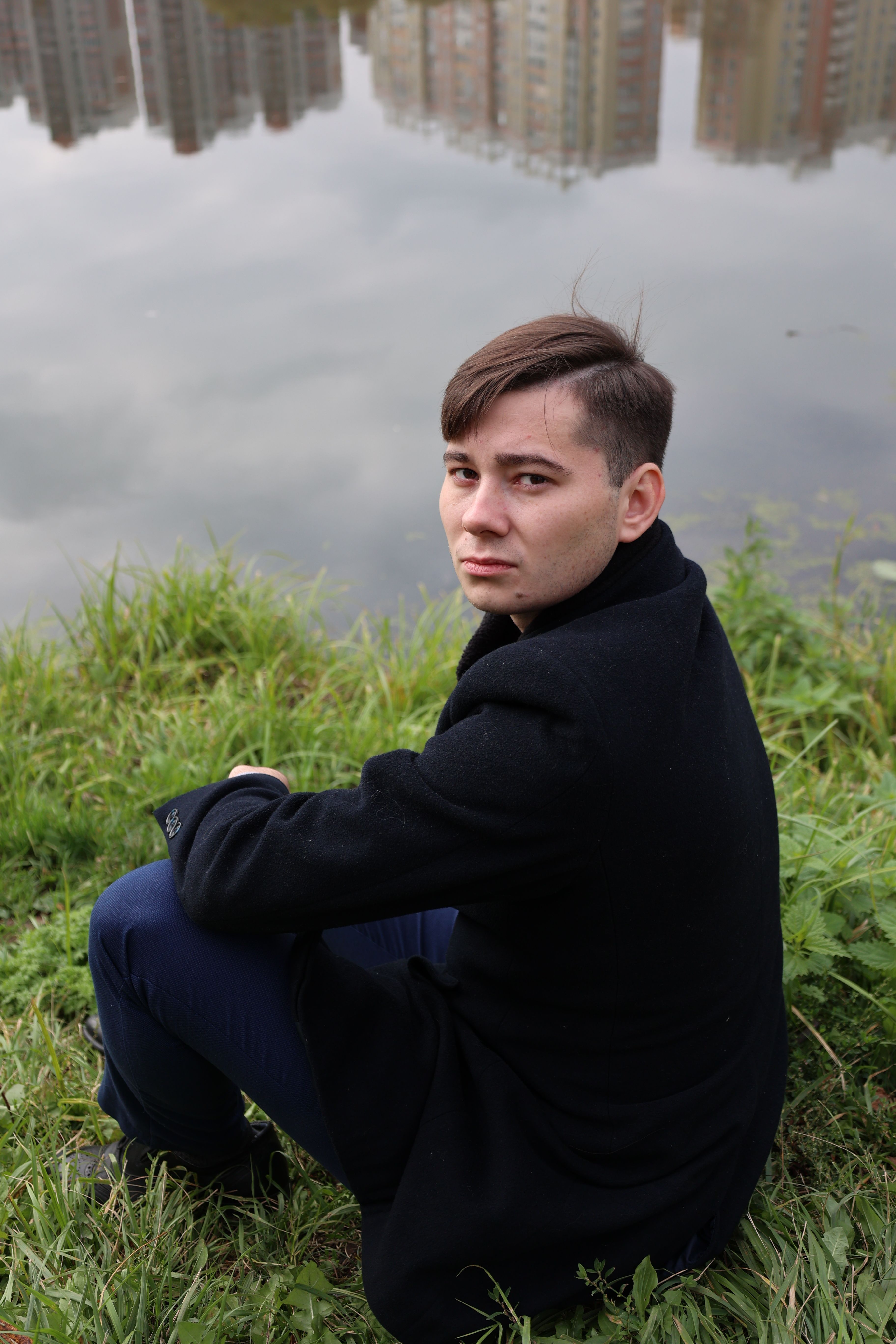 A young man in a suit jacket sits by a lakeside, gazing over his shoulder on an autumn day.