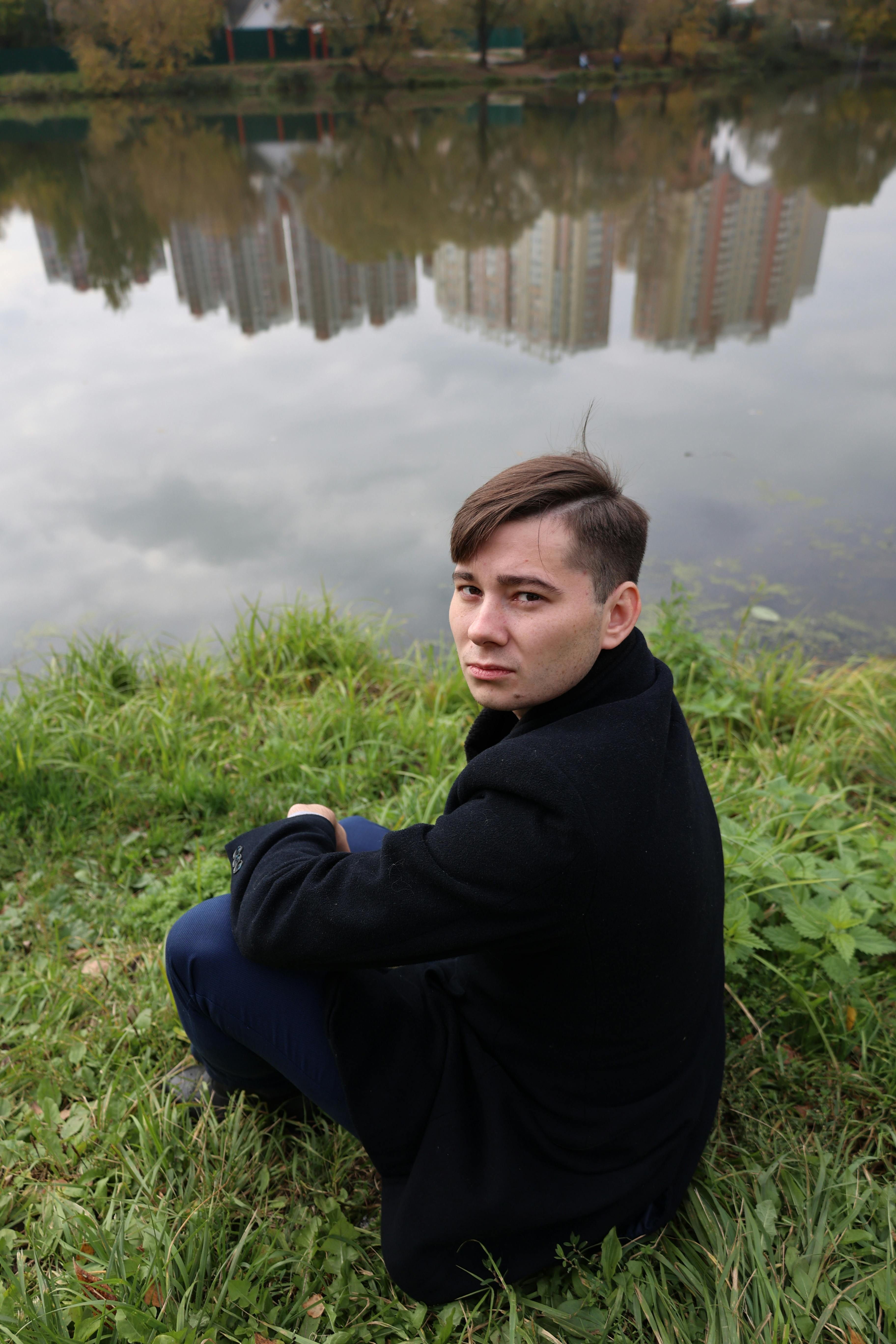 A young man in a black coat sits by a lake with city buildings reflected in the water.