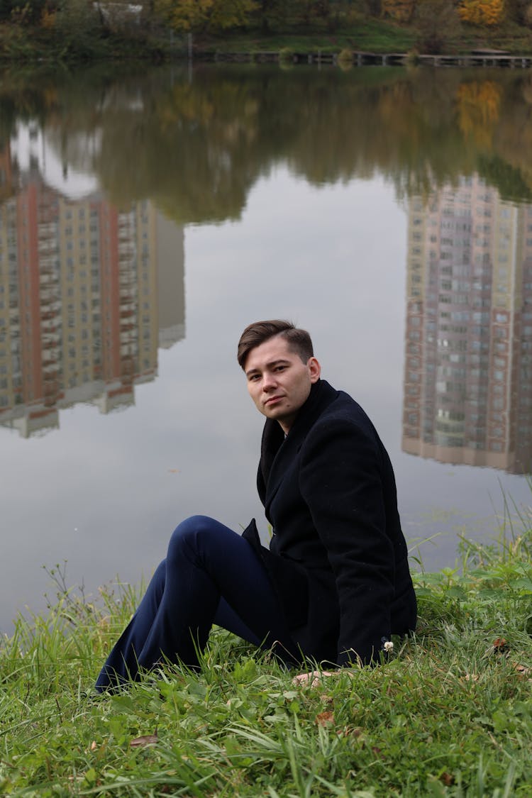 Young Man Sitting On Shore Of Lake In Estate