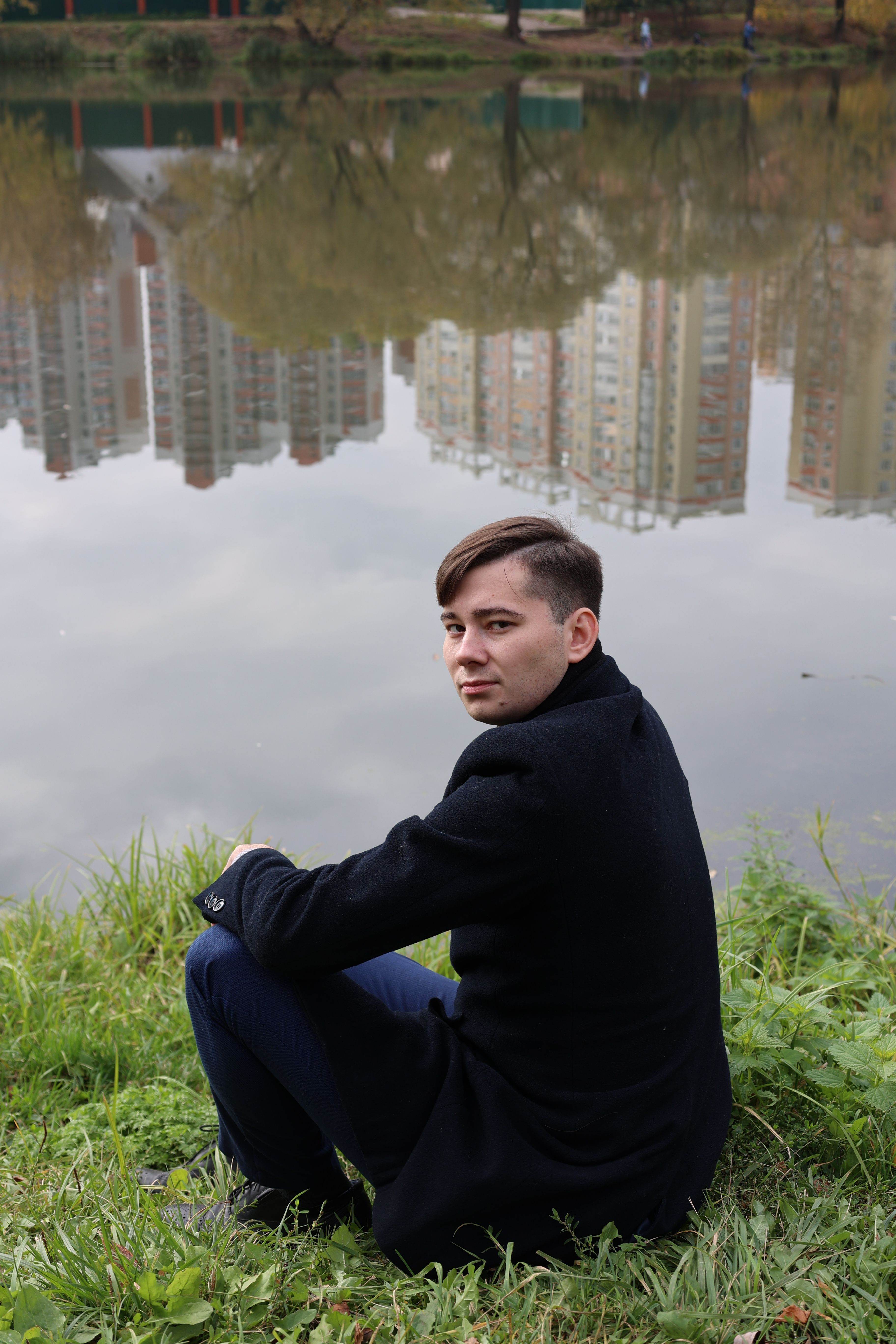 A young man in a dark coat sits by a lake reflecting urban buildings.