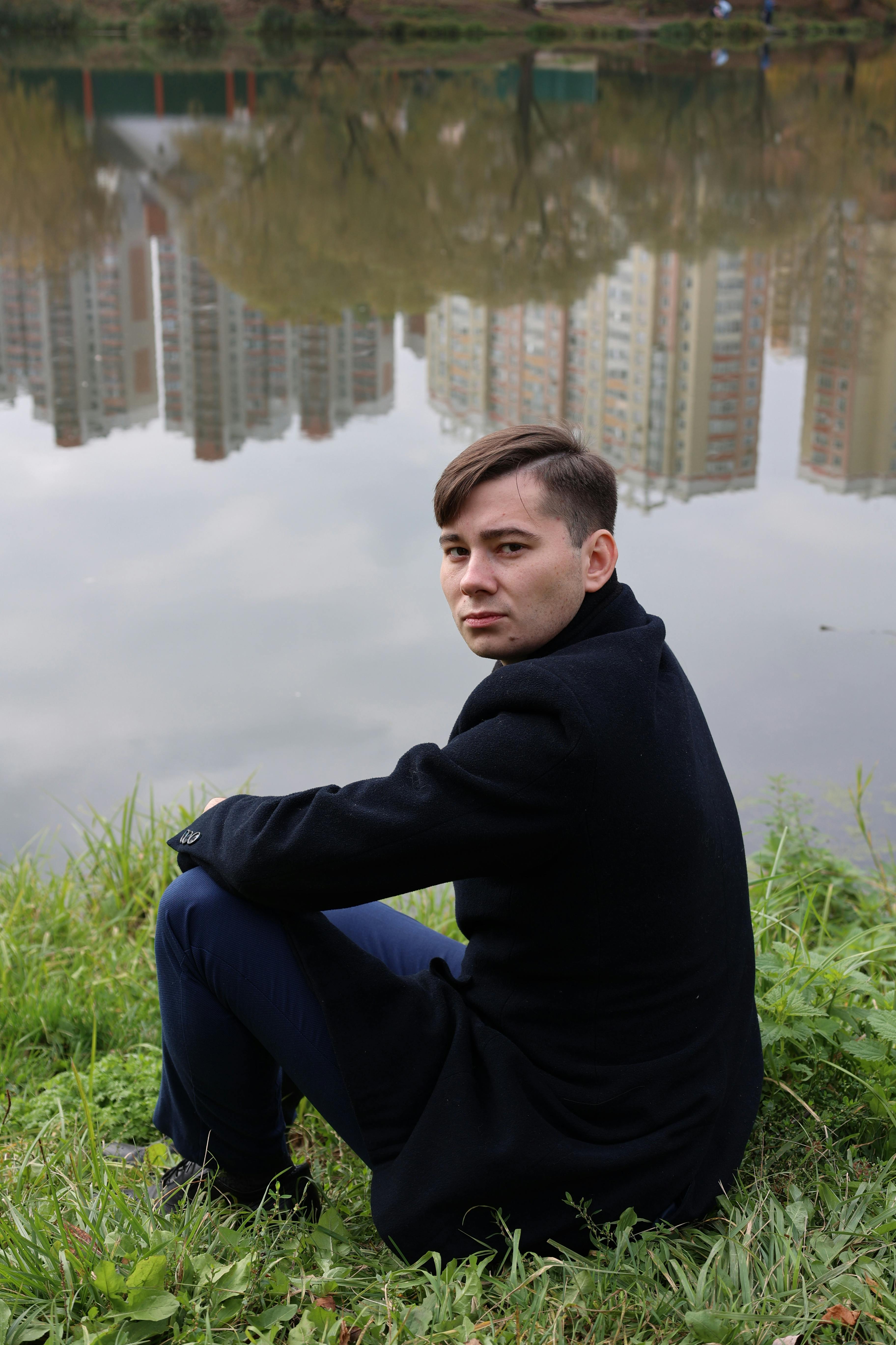 Young man in a black coat sitting by a lake with cityscape reflection.