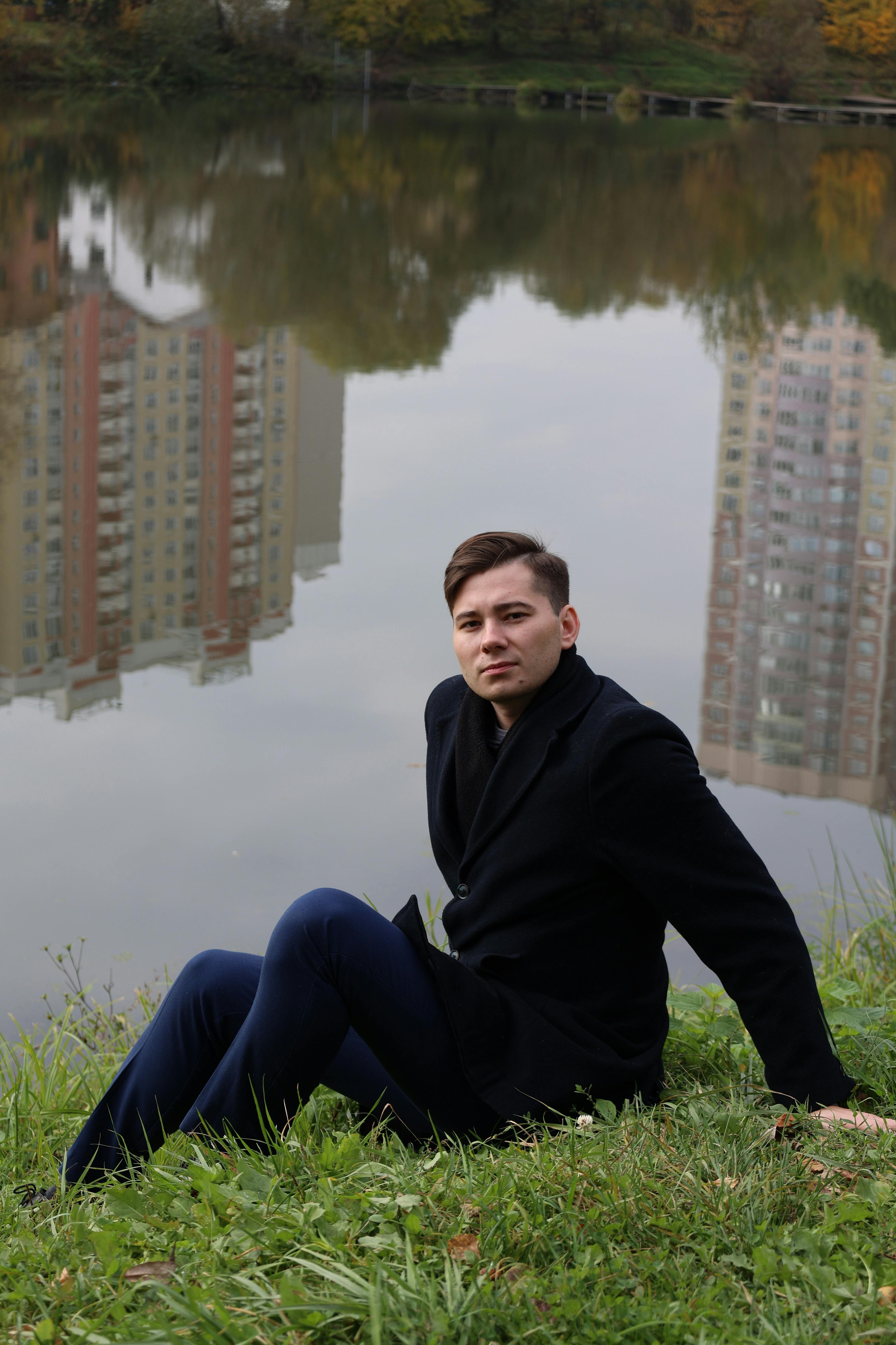 Caucasian man in a black coat sitting by a serene lake reflecting city buildings.