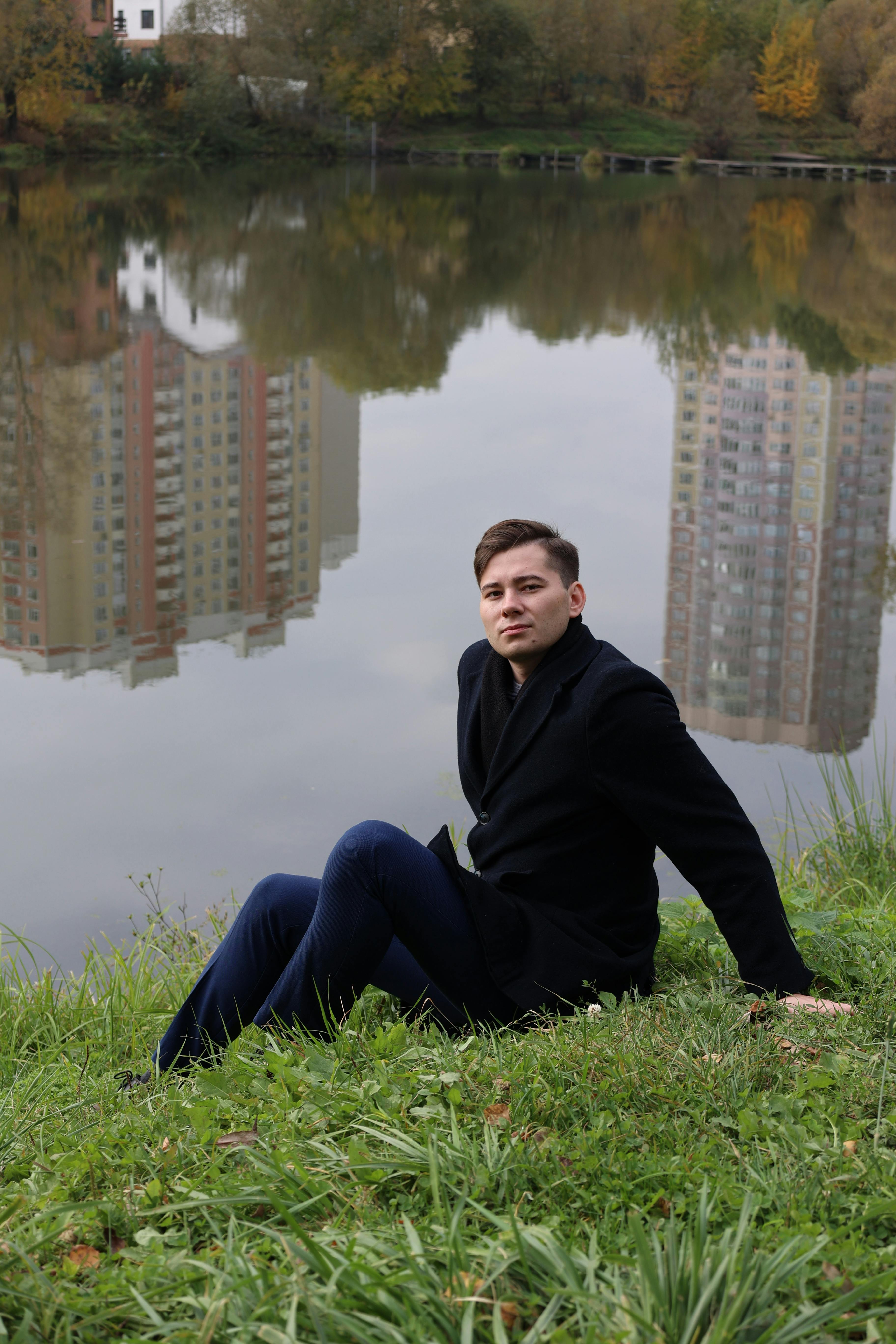Young man sitting by a lake with city buildings reflecting in the water.