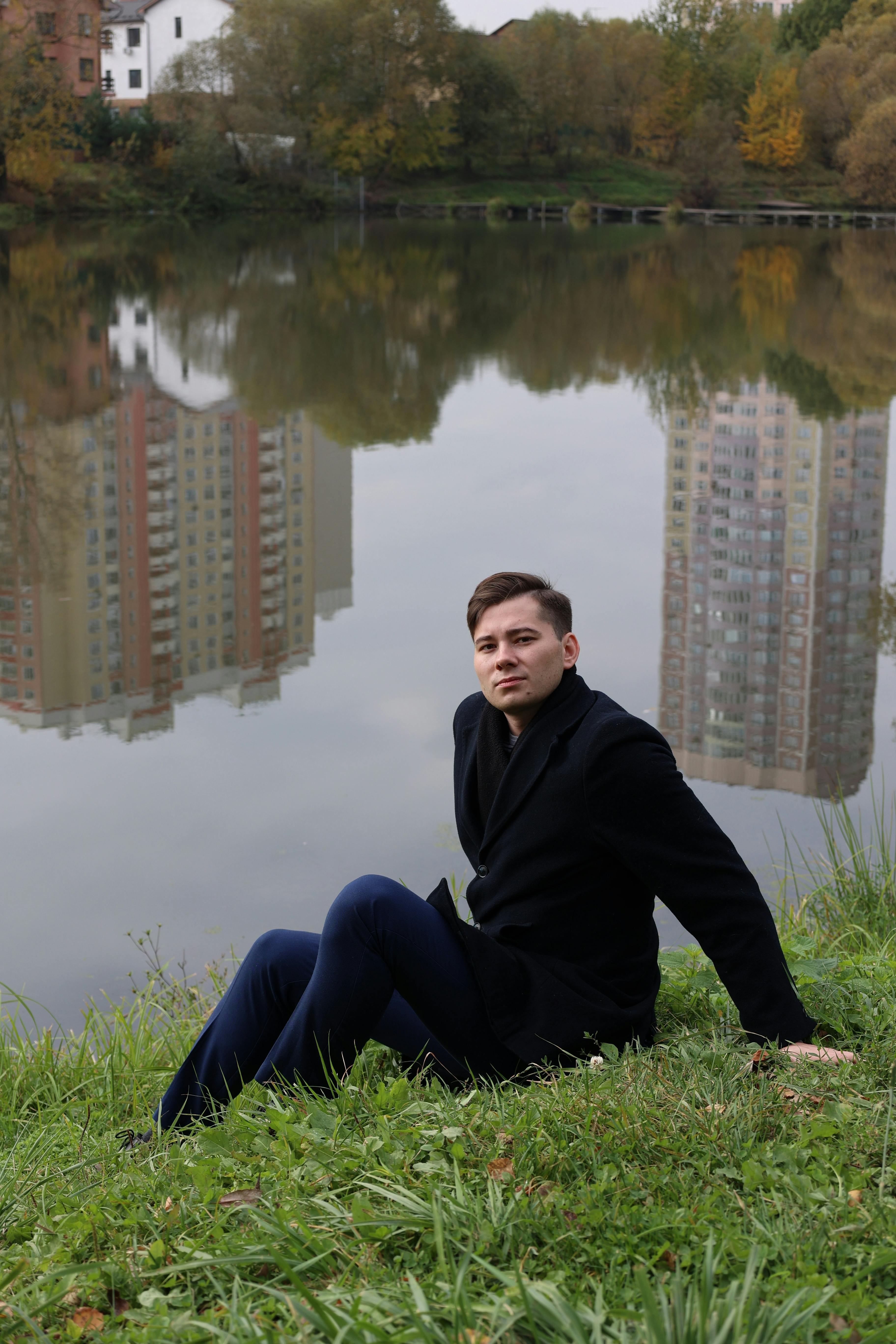 A man in a black coat sitting by a reflective lake with urban buildings in the background.