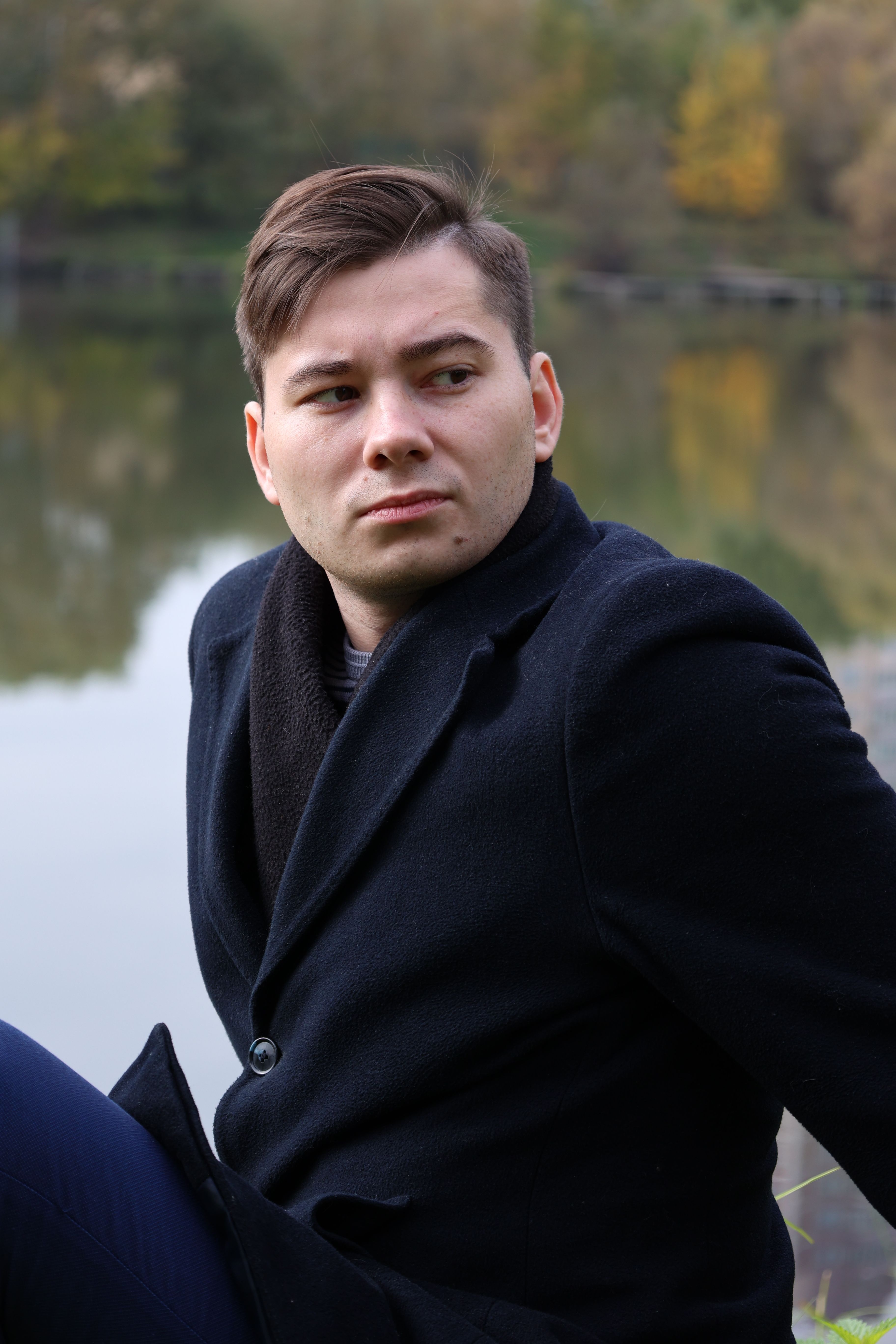 A thoughtful adult man in a black coat by a tranquil lake surrounded by autumn trees.