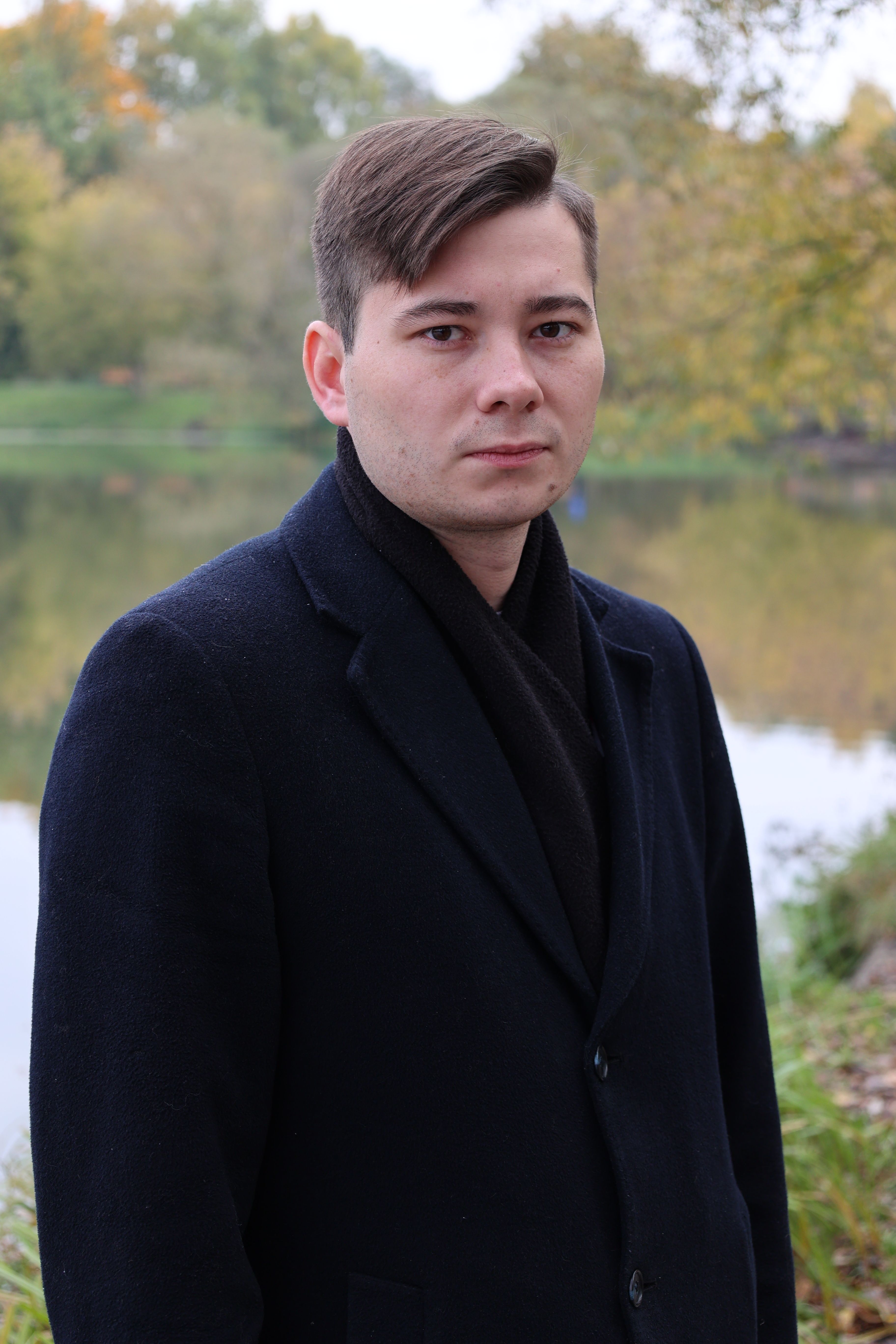 A serious man stands by a lake in autumn, wearing a dark coat.