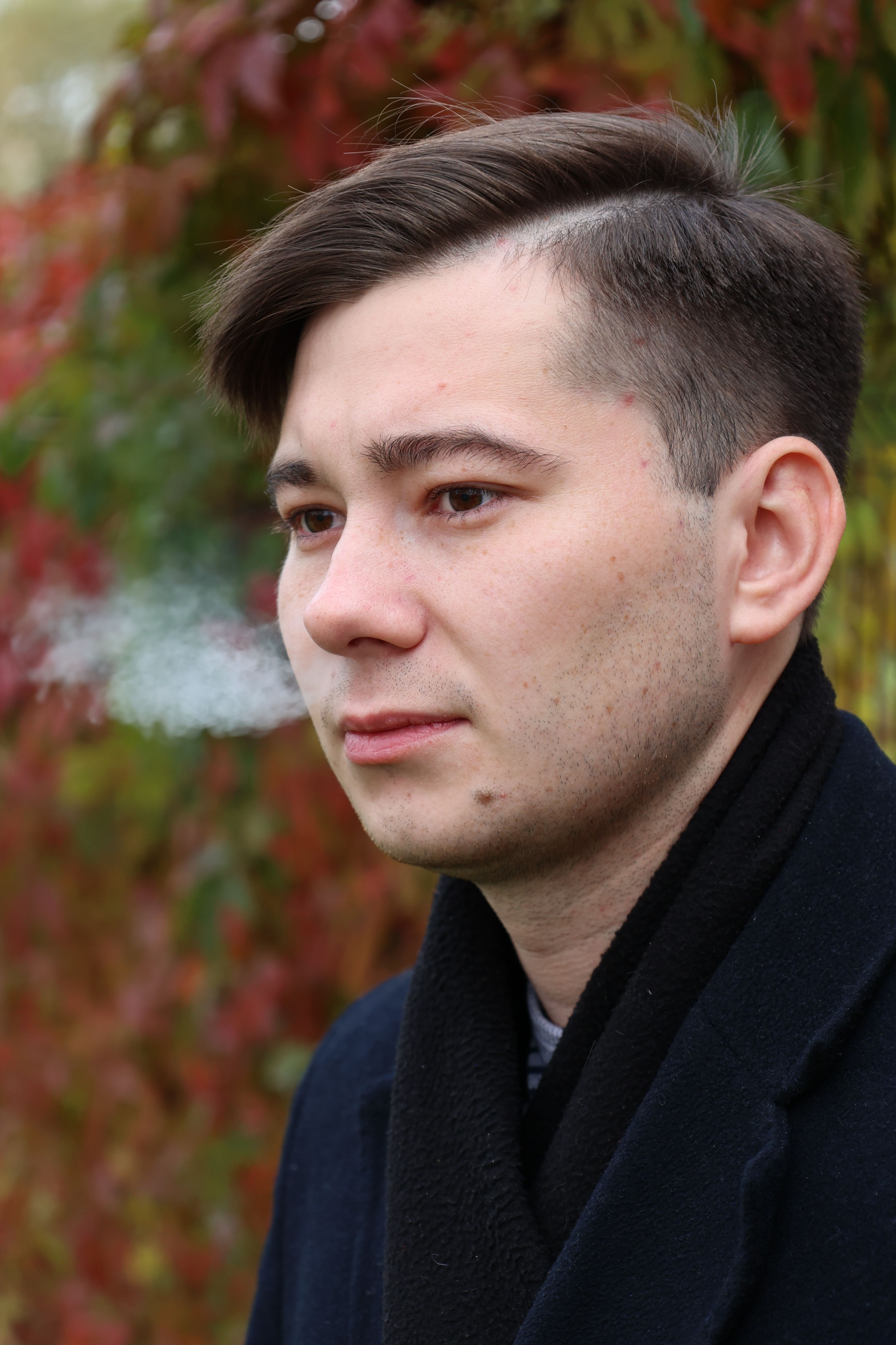 Close-up portrait of a young man smoking outdoors with autumn foliage background.