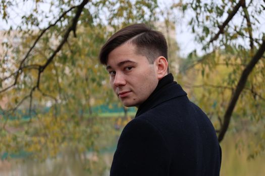 A man in a black coat glances back while standing in a serene autumn park.