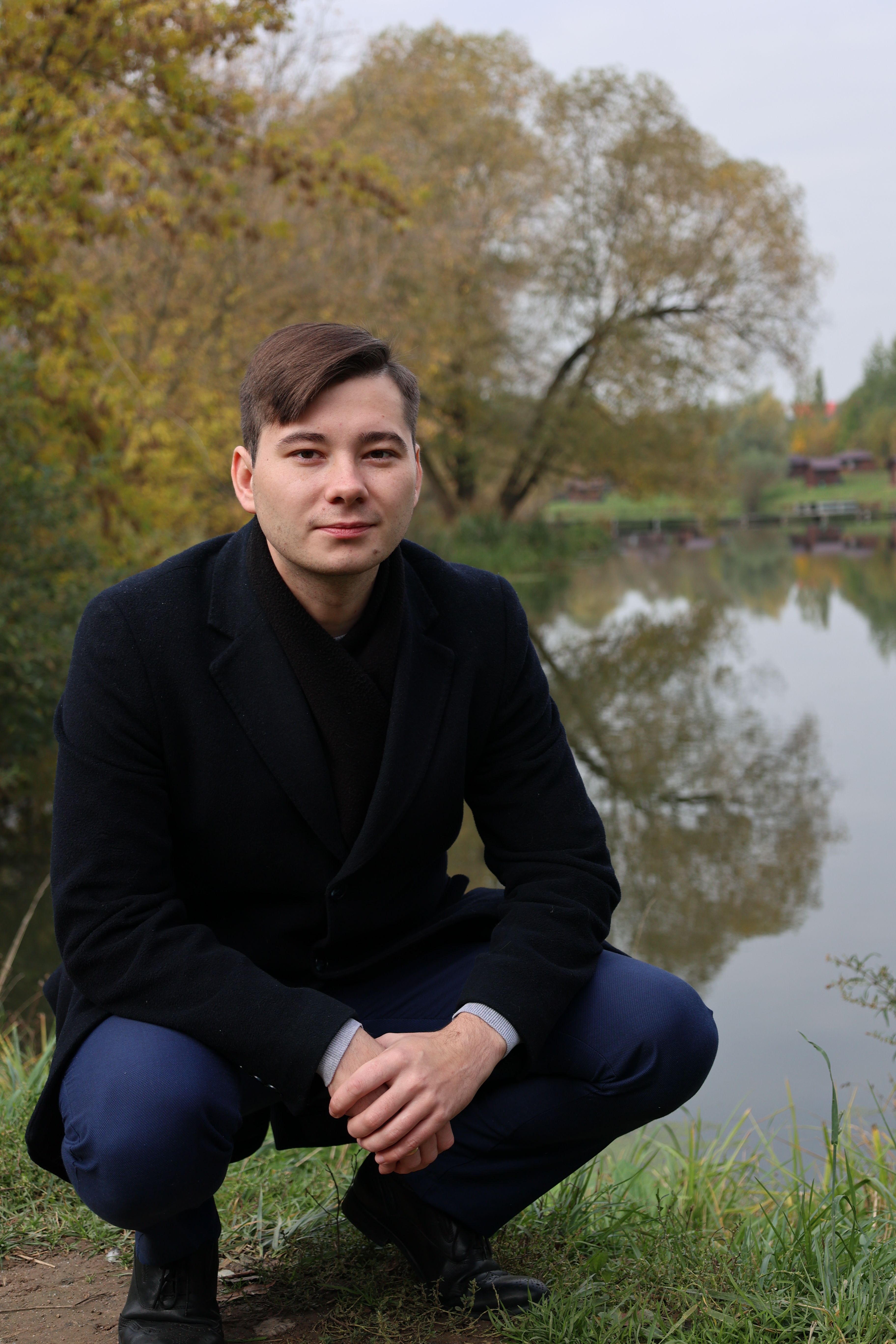 A man in a black coat sits by a tranquil lake surrounded by autumn trees.