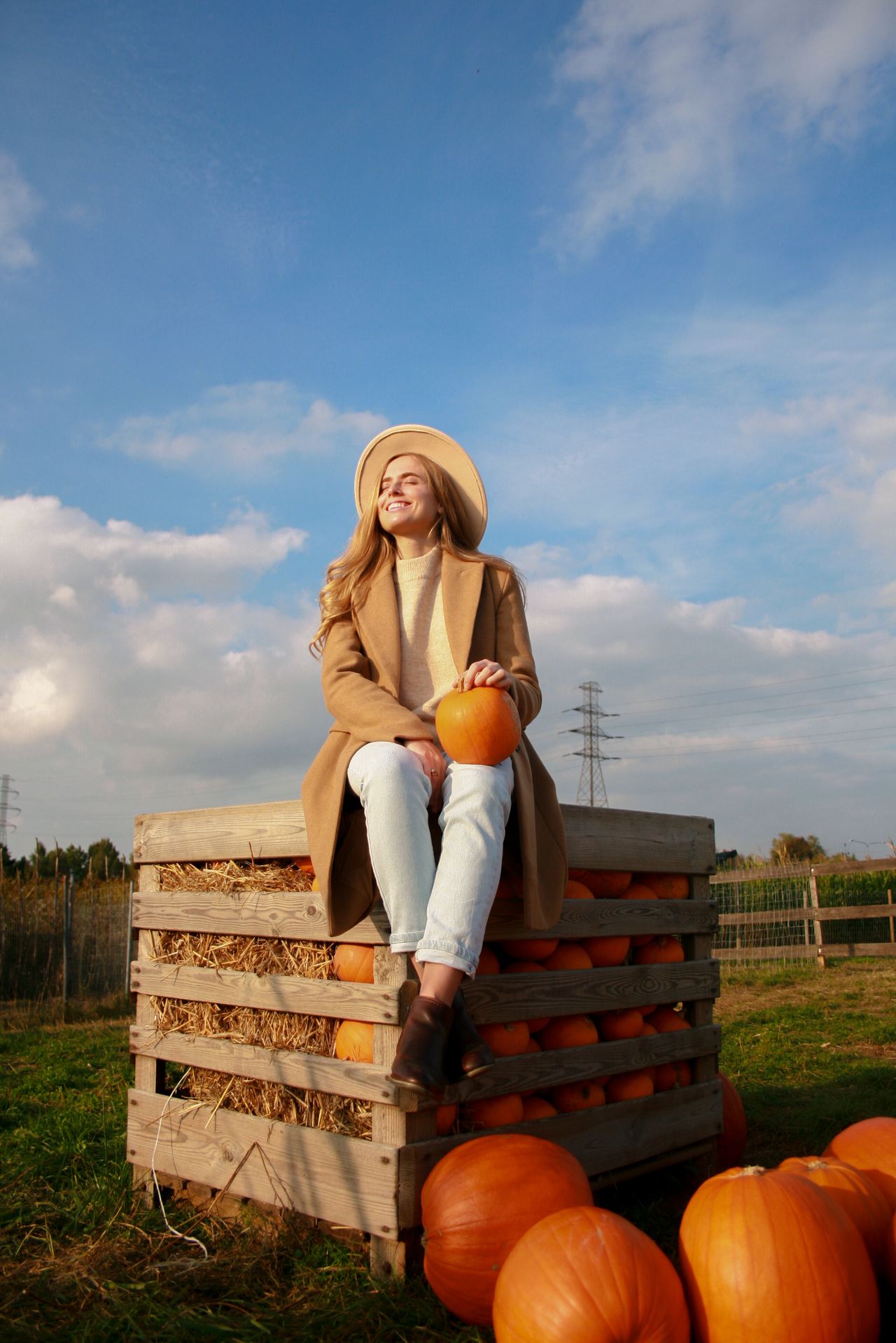 Woman in a camel coat, cream sweater and light jeans sitting on a wooden crate of pumpkins under a blue sky