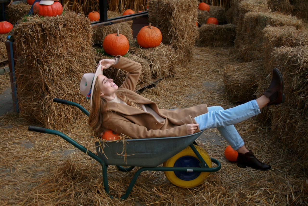 Woman in a camel coat and Chelsea boots laughing while reclining in a wheelbarrow among hay bales and pumpkins