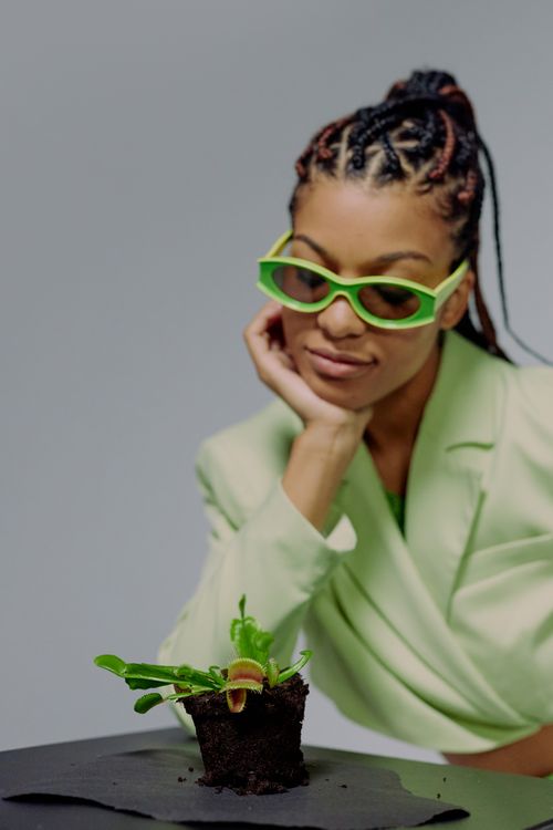 Free Elegant woman in green sunglasses sitting inside, observing a vibrant Venus flytrap plant on a table. Stock Photo