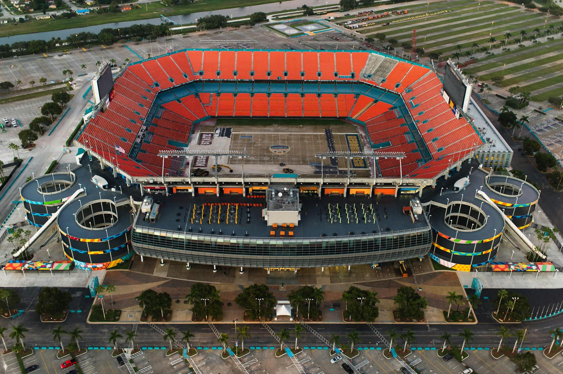 High-angle aerial shot of an empty sports stadium with orange seats and surrounding parking areas.