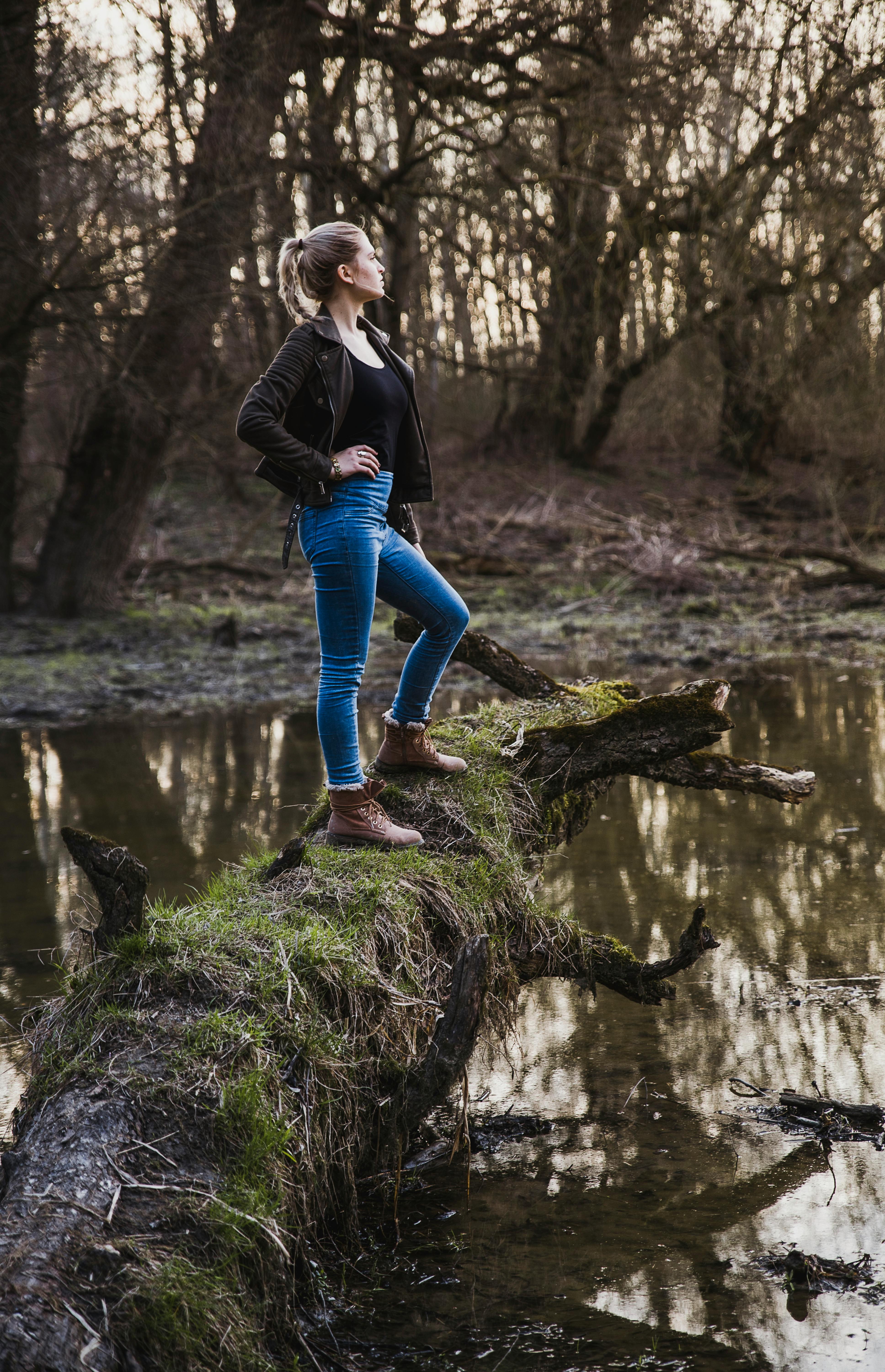 一位穿着牛仔裤和靴子的女人自信地站在宁静森林中倒映着河流的原木上