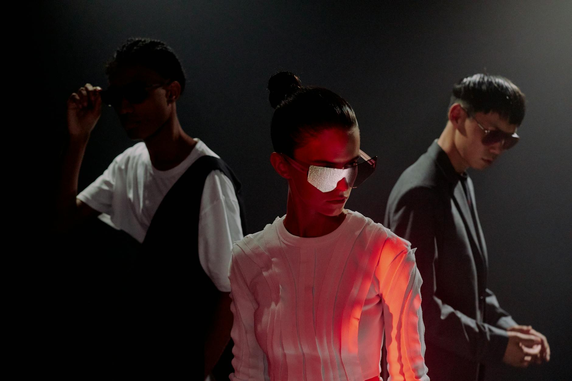 Three stylish individuals in a studio, showcasing futuristic eyewear fashion with dramatic lighting.
