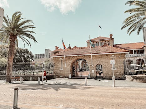 Historic Royal Mint building with flags and palm trees under a bright sky.