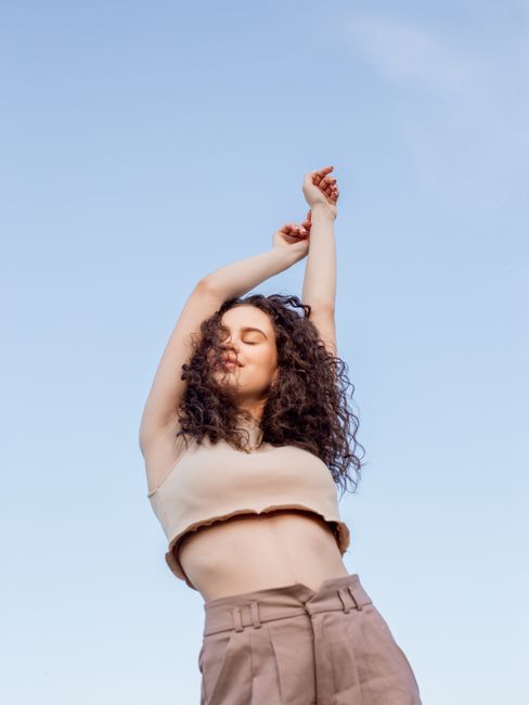 A young woman with curly hair joyfully stretches her arms under the bright blue sky, embodying freedom and serenity.