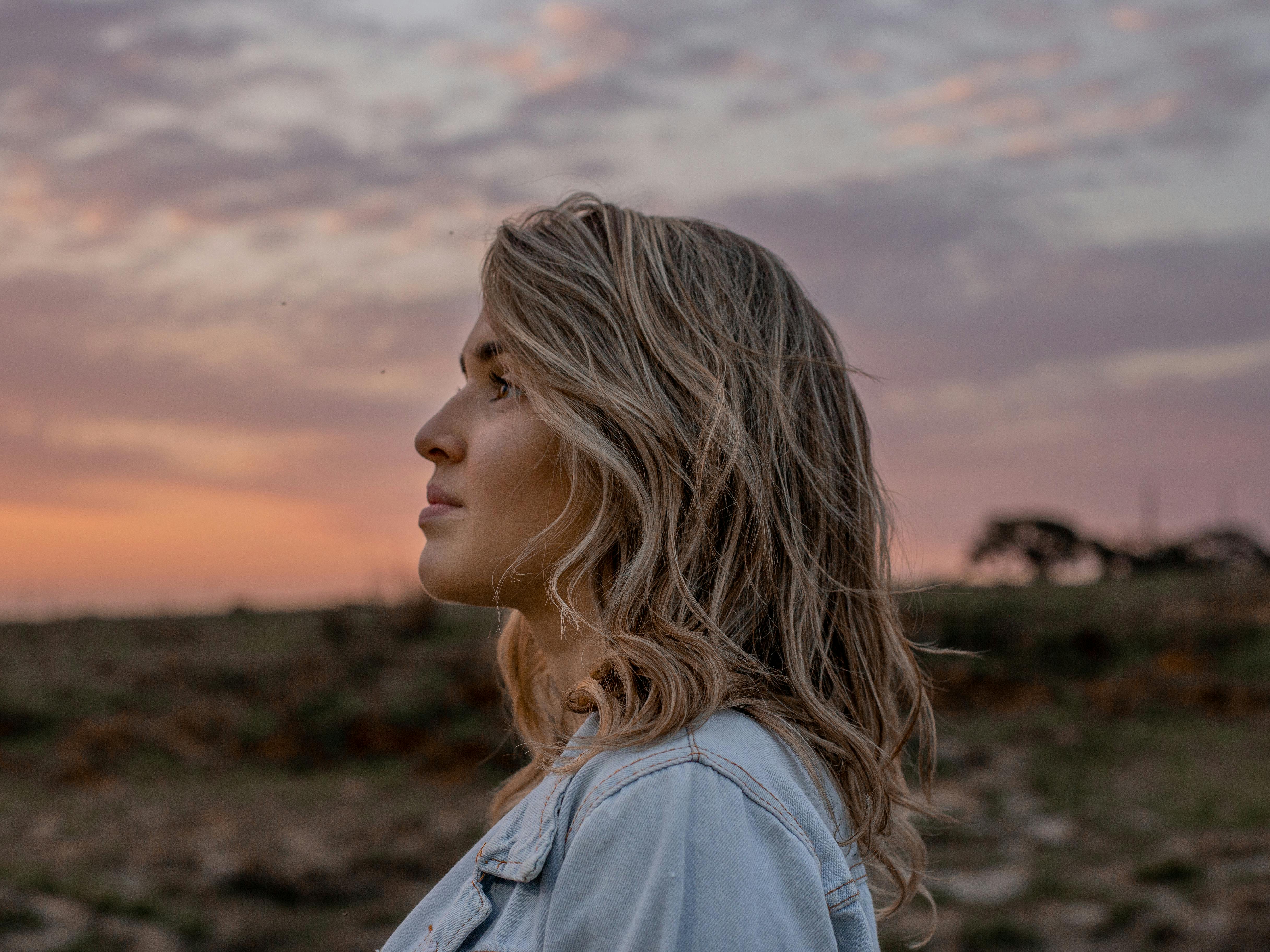 Woman with wavy blonde hair gazes thoughtfully at a serene sunset in the countryside, wearing a denim jacket. Sky hues softly blend pink and orange.