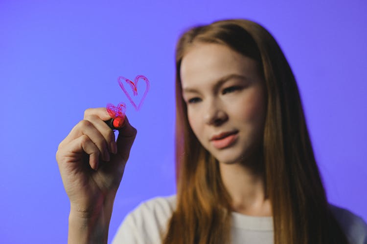 Girl Drawing Heart On Glass Against Blue Background