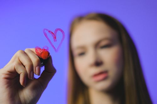 Free Teen girl with long brown hair draws red heart on glass against blue background. Stock Photo
