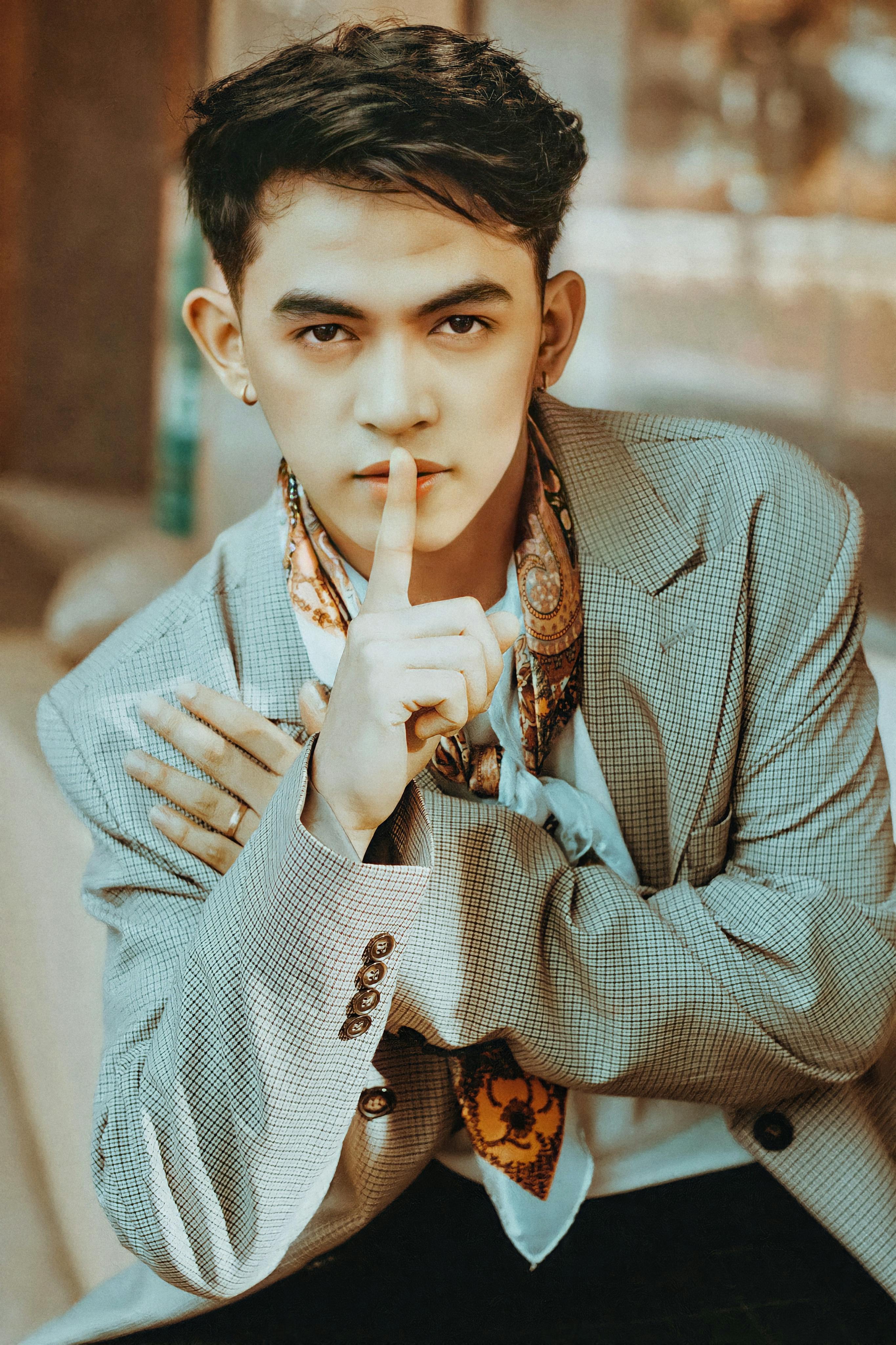 Young man in a checkered blazer making a hush gesture while posing confidently.