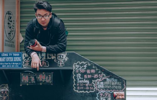 Free Young man with camera poses beside a menu board in an urban outdoor scene, displaying casual fashion. Stock Photo