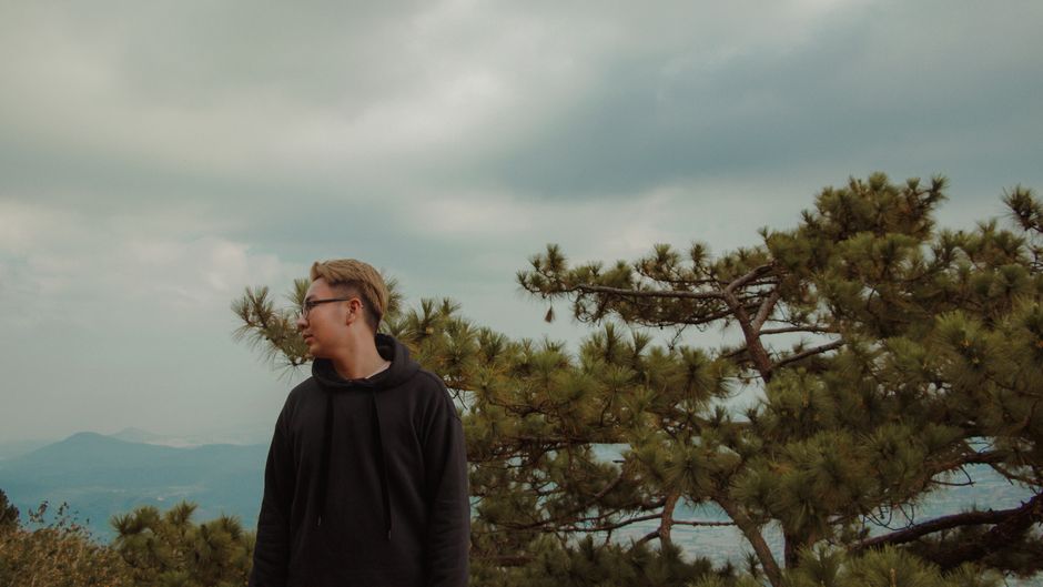 Profile of a young man in glasses at a park with trees and a cloudy sky.