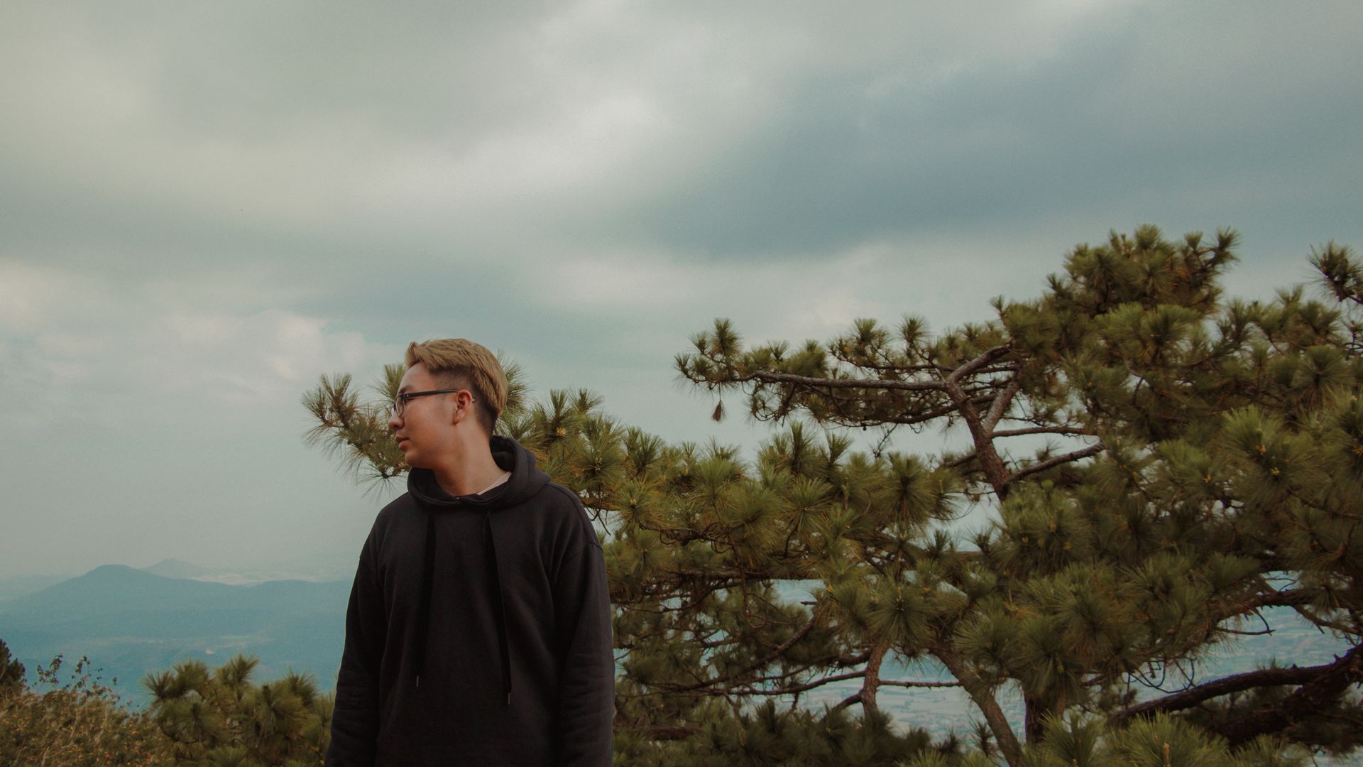Profile of a young man in glasses at a park with trees and a cloudy sky.