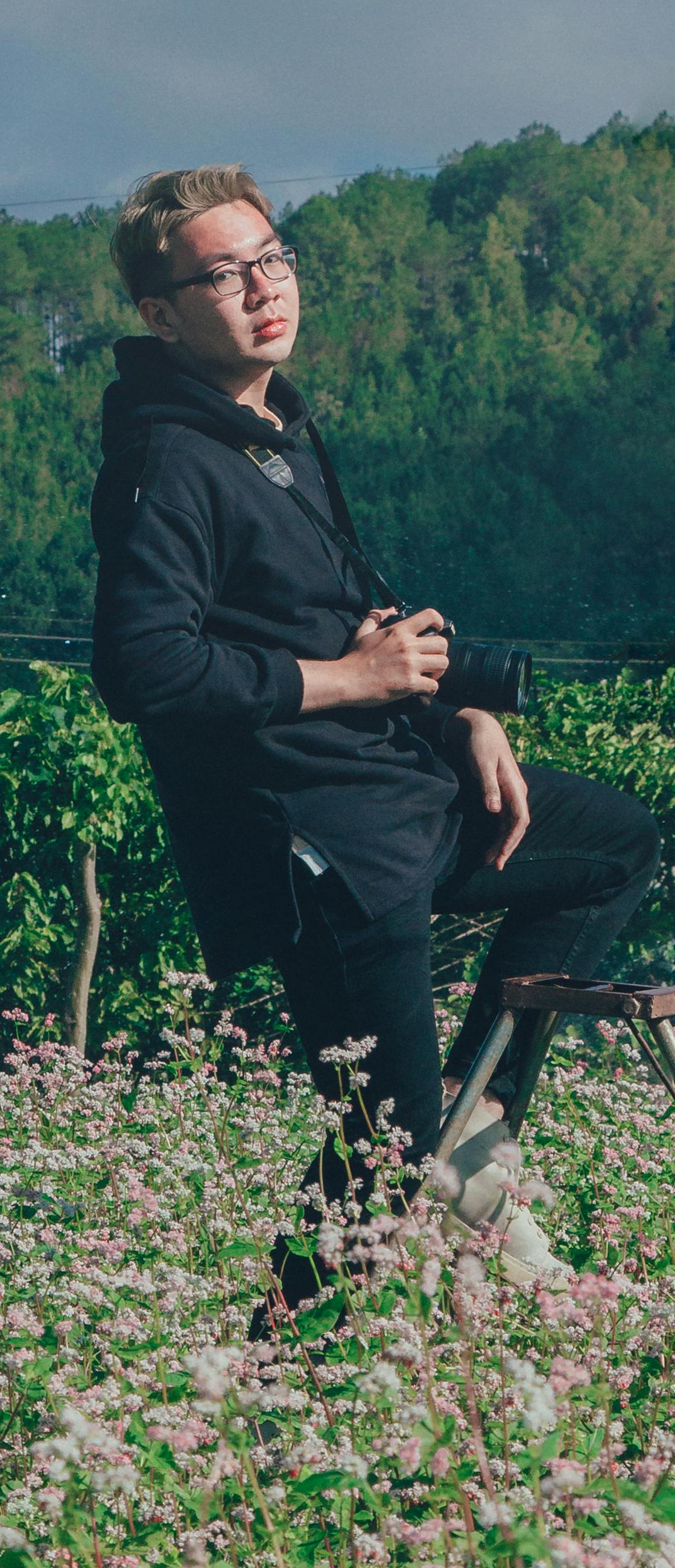 Young man in a garden holding a camera, surrounded by lush green flora.