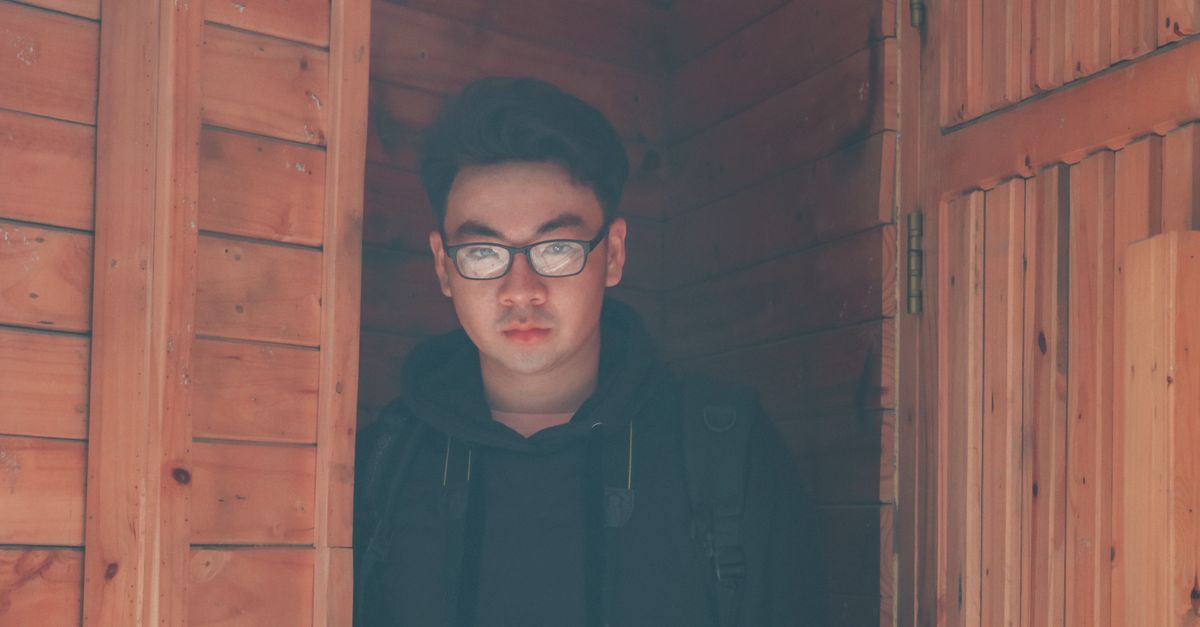 Um jovem usando óculos está parado em uma porta de madeira, capturado à luz natural do dia.