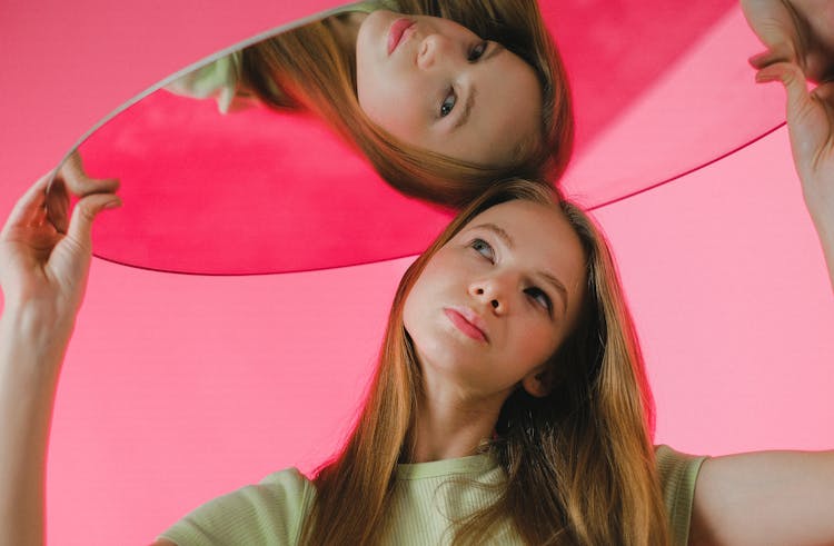 A Teenager Holding A Mirror Over Her Head and Looking At Her Reflection 