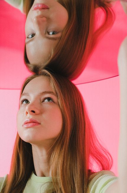 A thoughtful teenage girl with long brown hair gazing into her reflection on a pink background.
