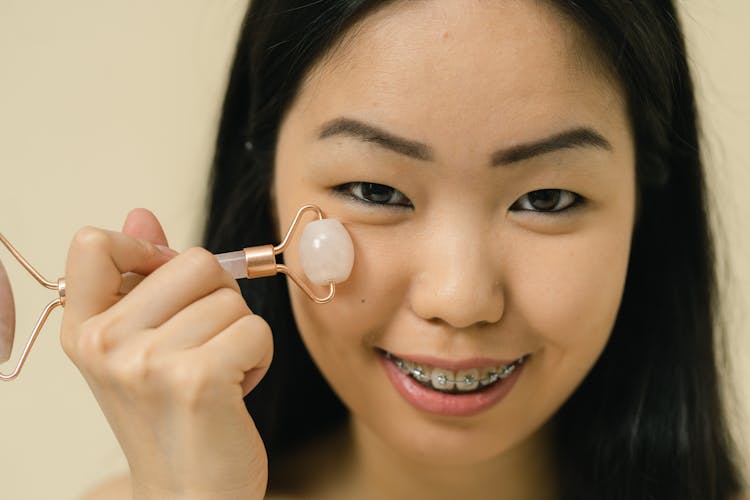 An Asian Woman Looking At A Camera And Using A gua sha roller 