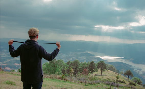 A man in a hoodie overlooks a scenic mountainous landscape with sunrays peeping through clouds.