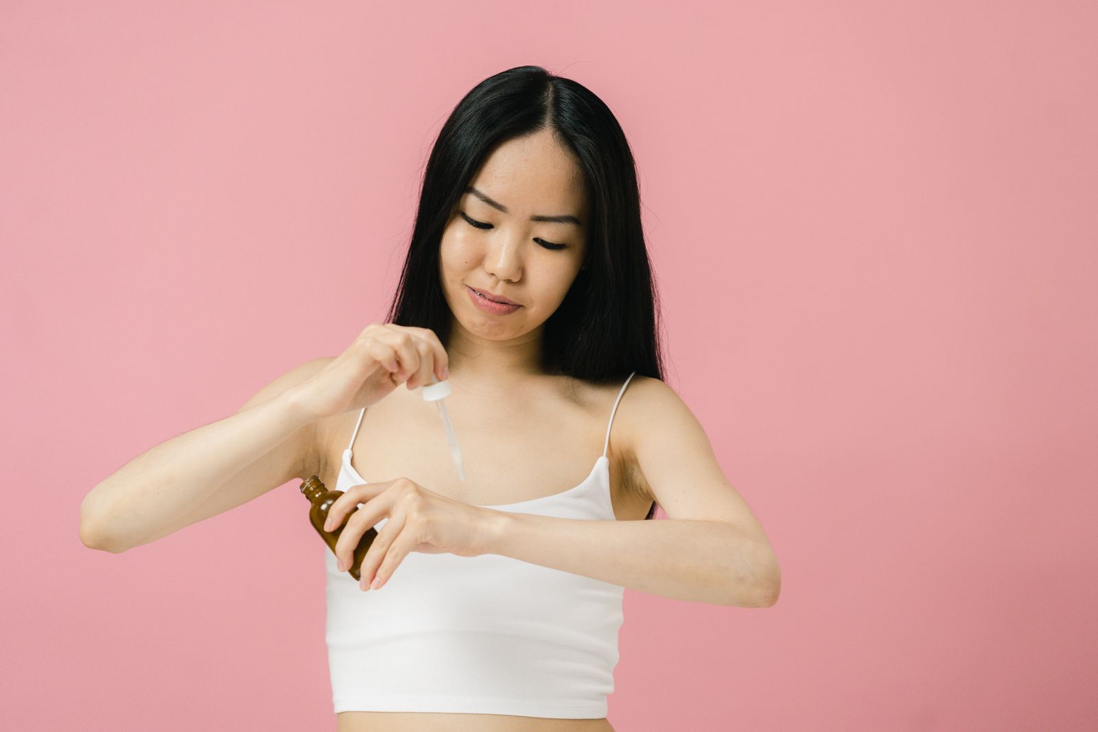 Woman with long dark hair holding an amber serum bottle and dropper against a pink background