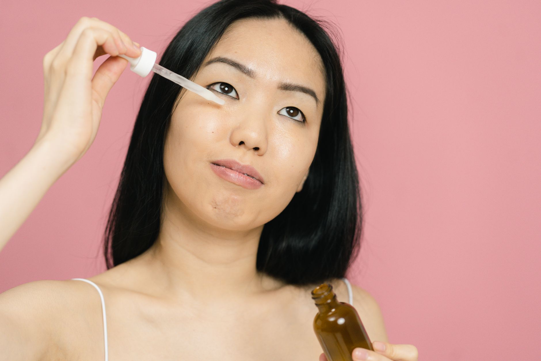 How to Use Peptides for Anti-Aging in the US 1 A woman applying facial serum with a dropper for skin care on a pink background.