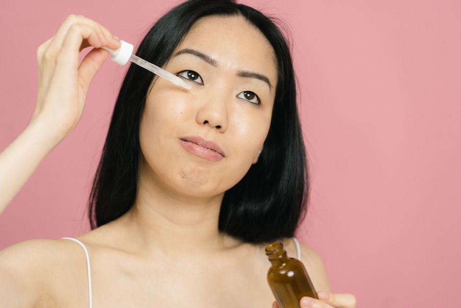A woman applying facial serum with a dropper for skin care on a pink background.