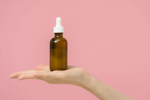 A brown dropper bottle rests on a hand against a pink background, perfect for skincare concepts.