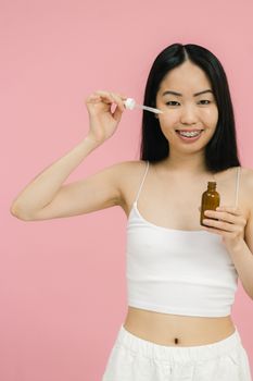 Young woman in tank top applying serum to face with dropper, smiling.
