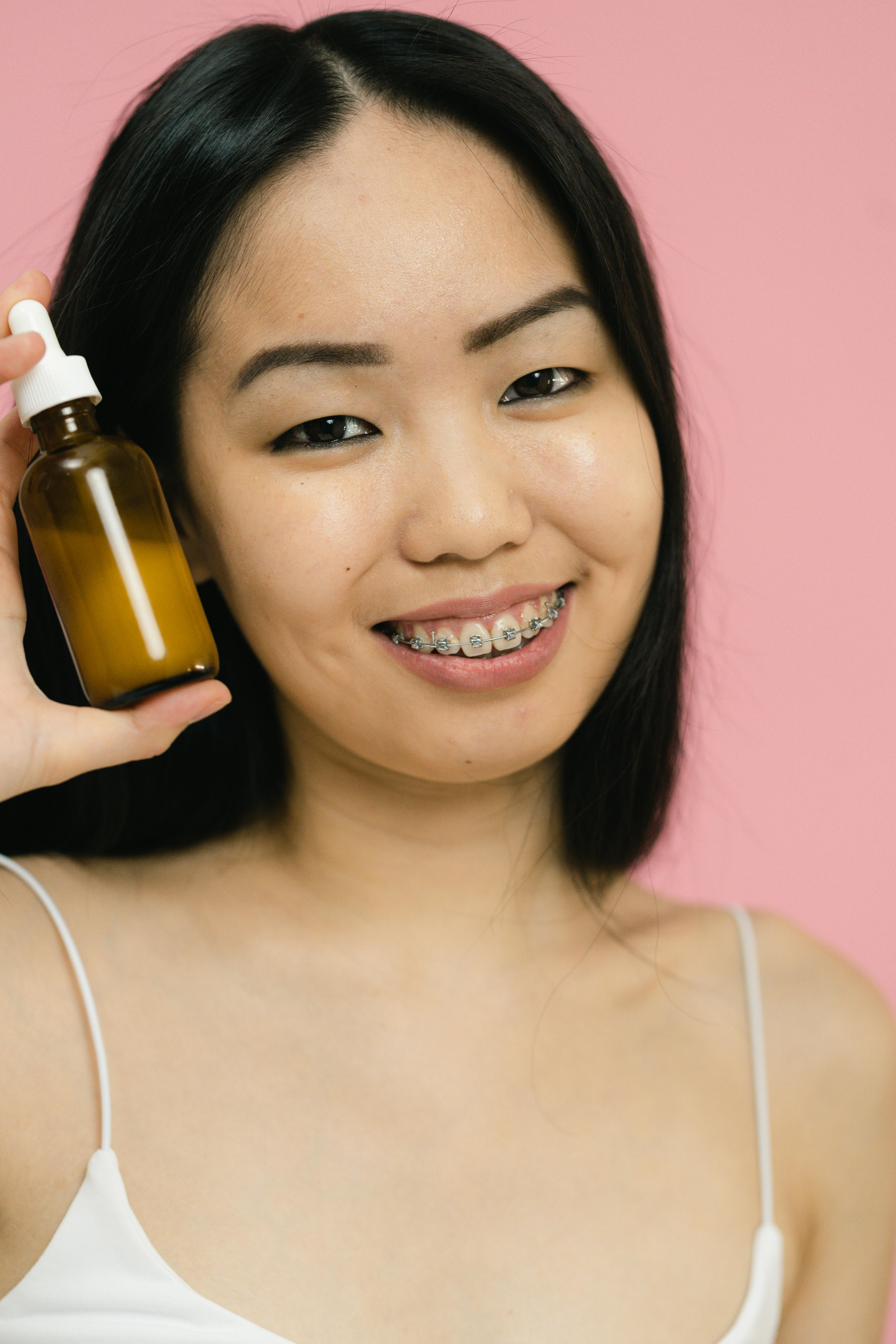 Smiling woman with braces holding a cosmetic bottle against a pink background.
